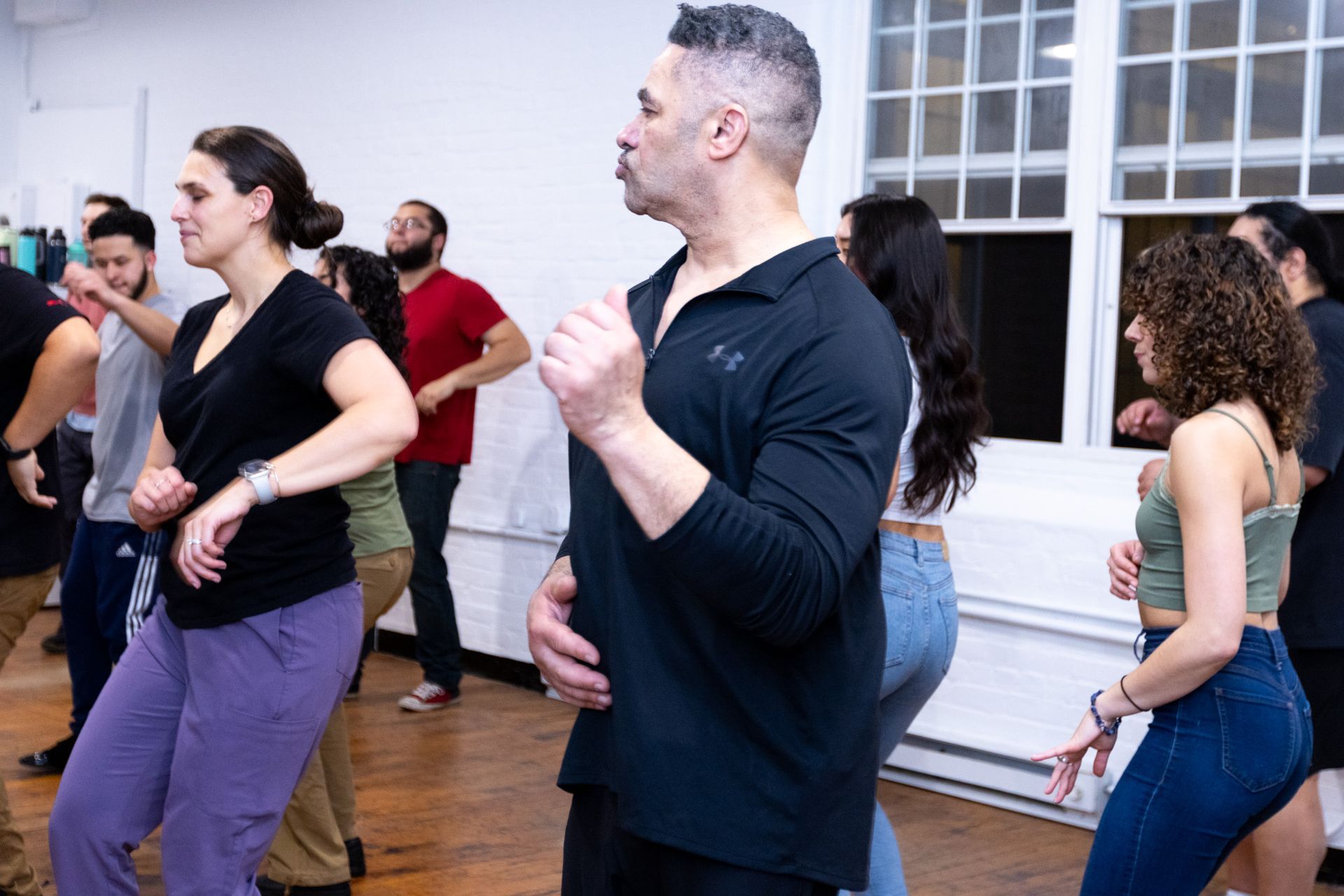 A group of younger and older men and women practice footwork choreography during a Bachata class.