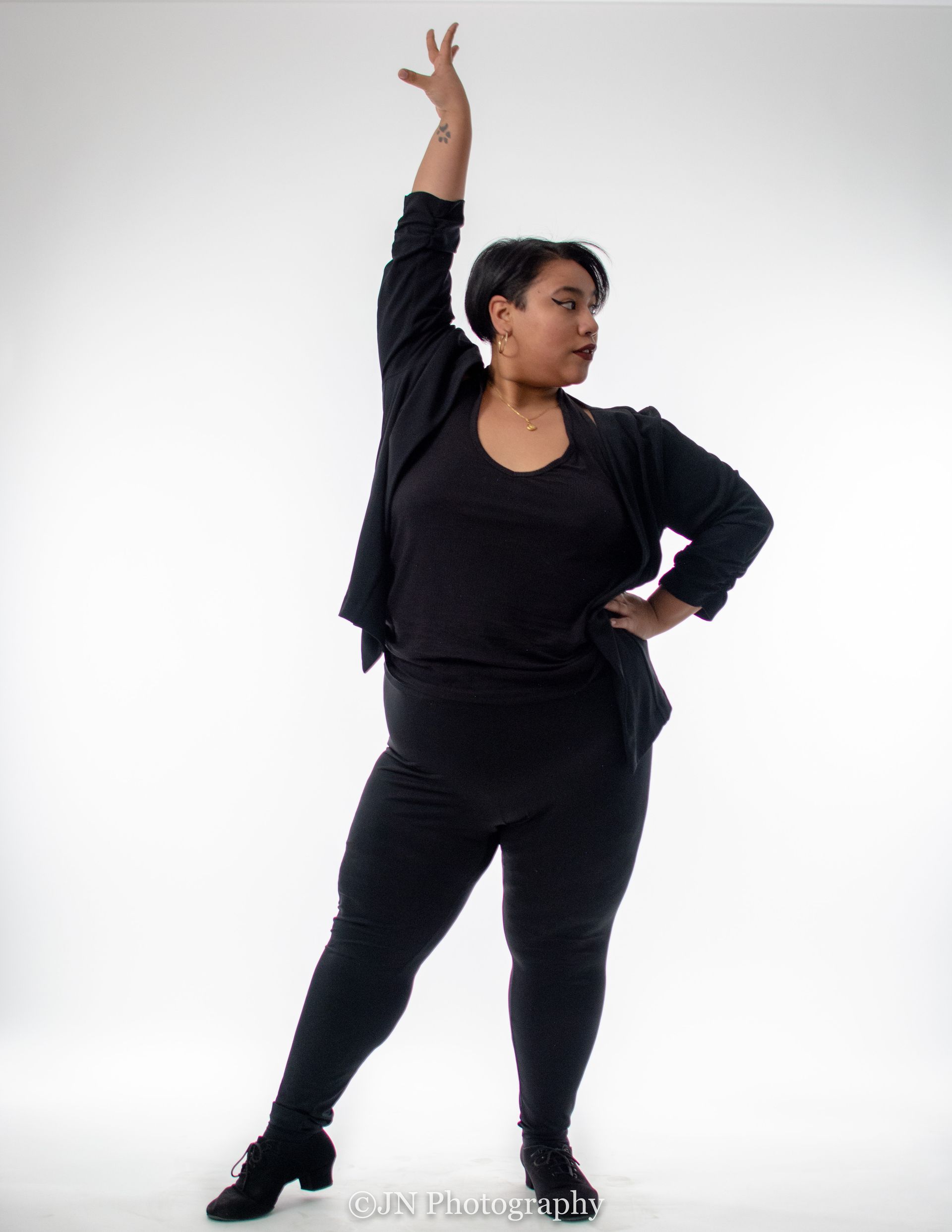 Dance instructor Kristy Berdugo wearing all black poses with a hand on her hip, an arm up in the air, and a toe pointed out to the side. 