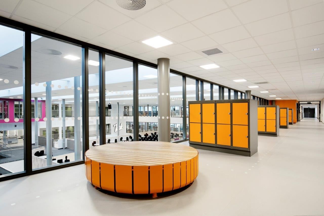 Moderne skolegang med store vinduer, oransje skap og en rund sittegruppe.