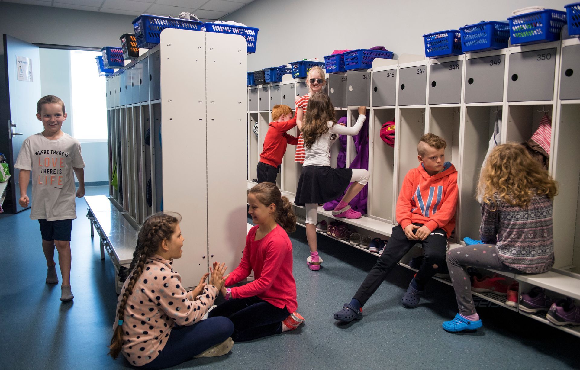 Barn i en skolegang med skap, samhandler. Noen sitter, noen står. Lyst og muntert miljø.