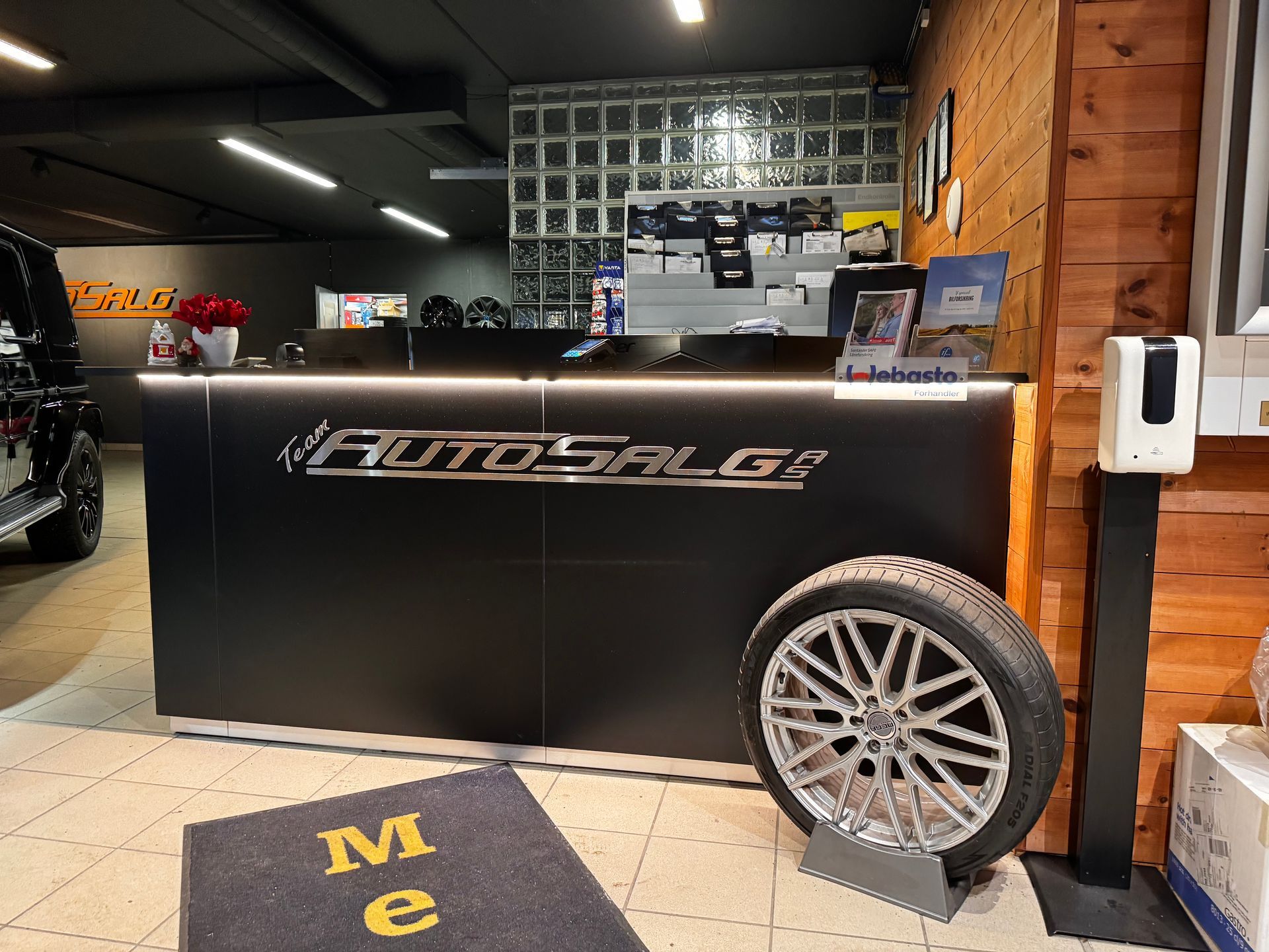 Svart resepsjon med «Auto Salg»-logo, et dekk og en velkomstmatte inne i en bilforhandler.
