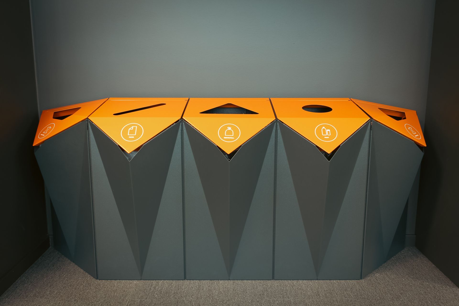 Resirkuleringsstasjon med fem søppelbøtter, oransje lokk og geometriske grå sider. 