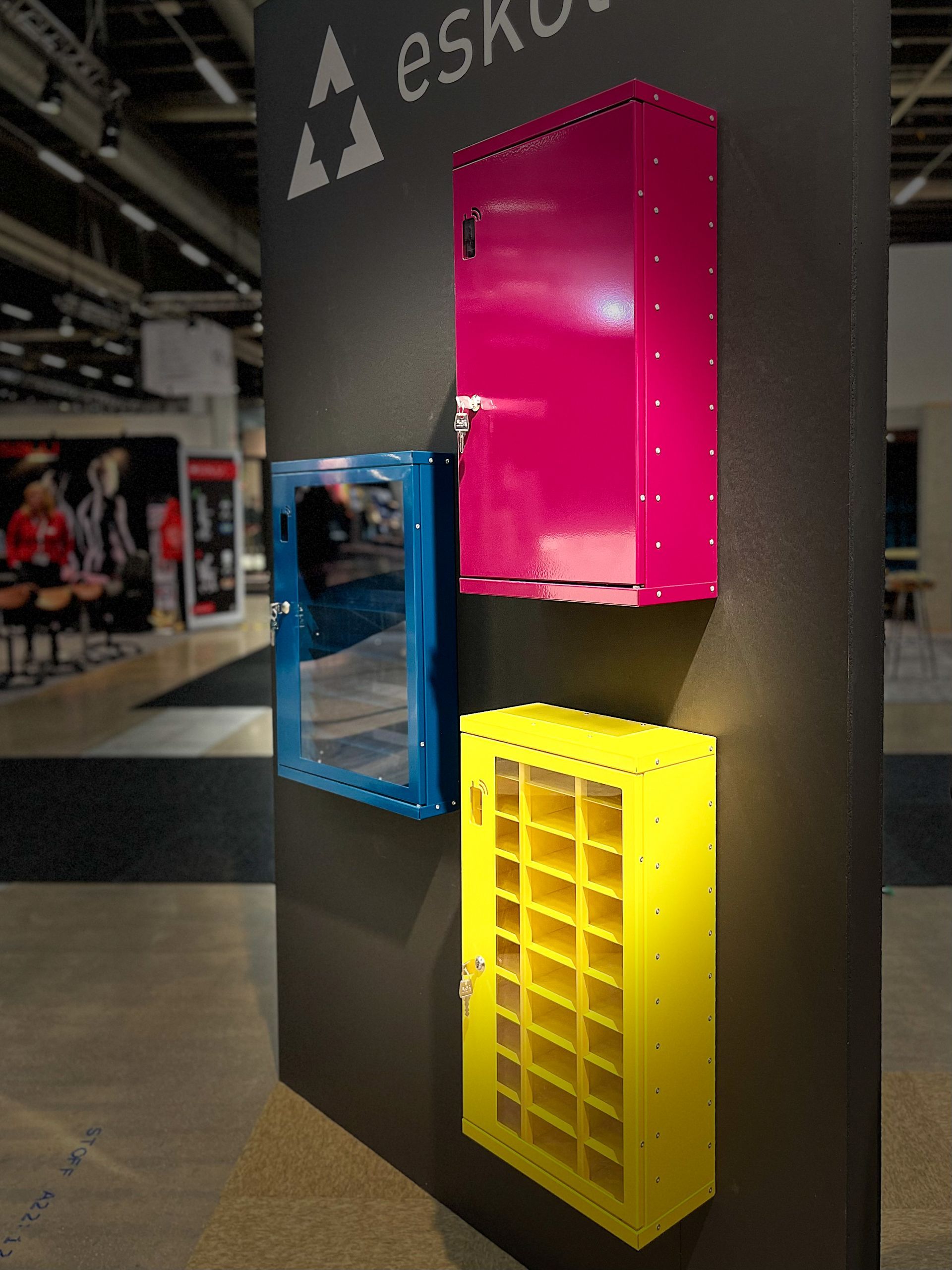 Tre fargerike veggmonterte skap: blå med speil, rosa og gule med små oppbevaringsskap, på en mørkegrå søyle.