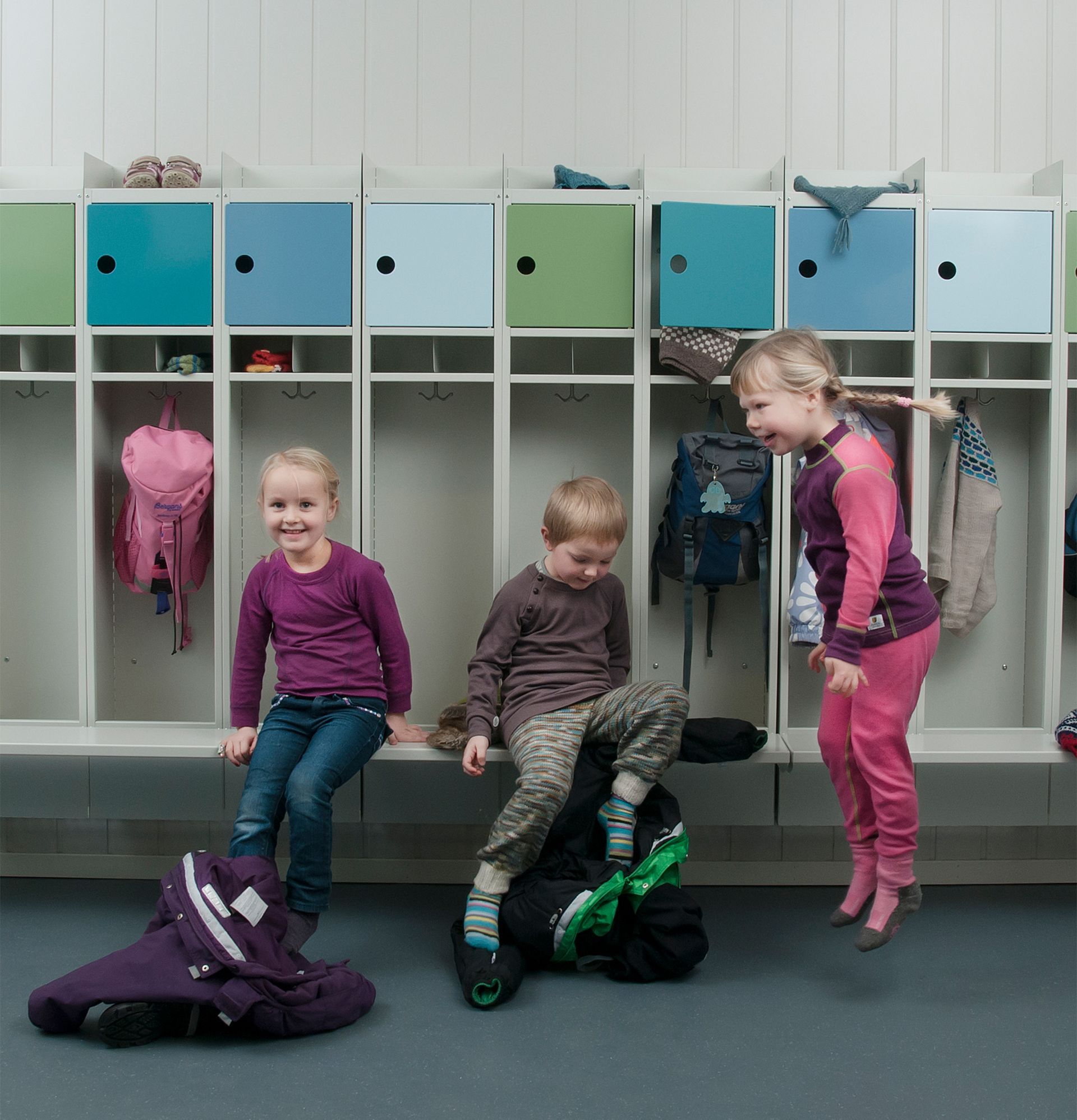Tre barn i en skolegang: to sitter, ett hopper. Fargerike skap i bakgrunnen.
