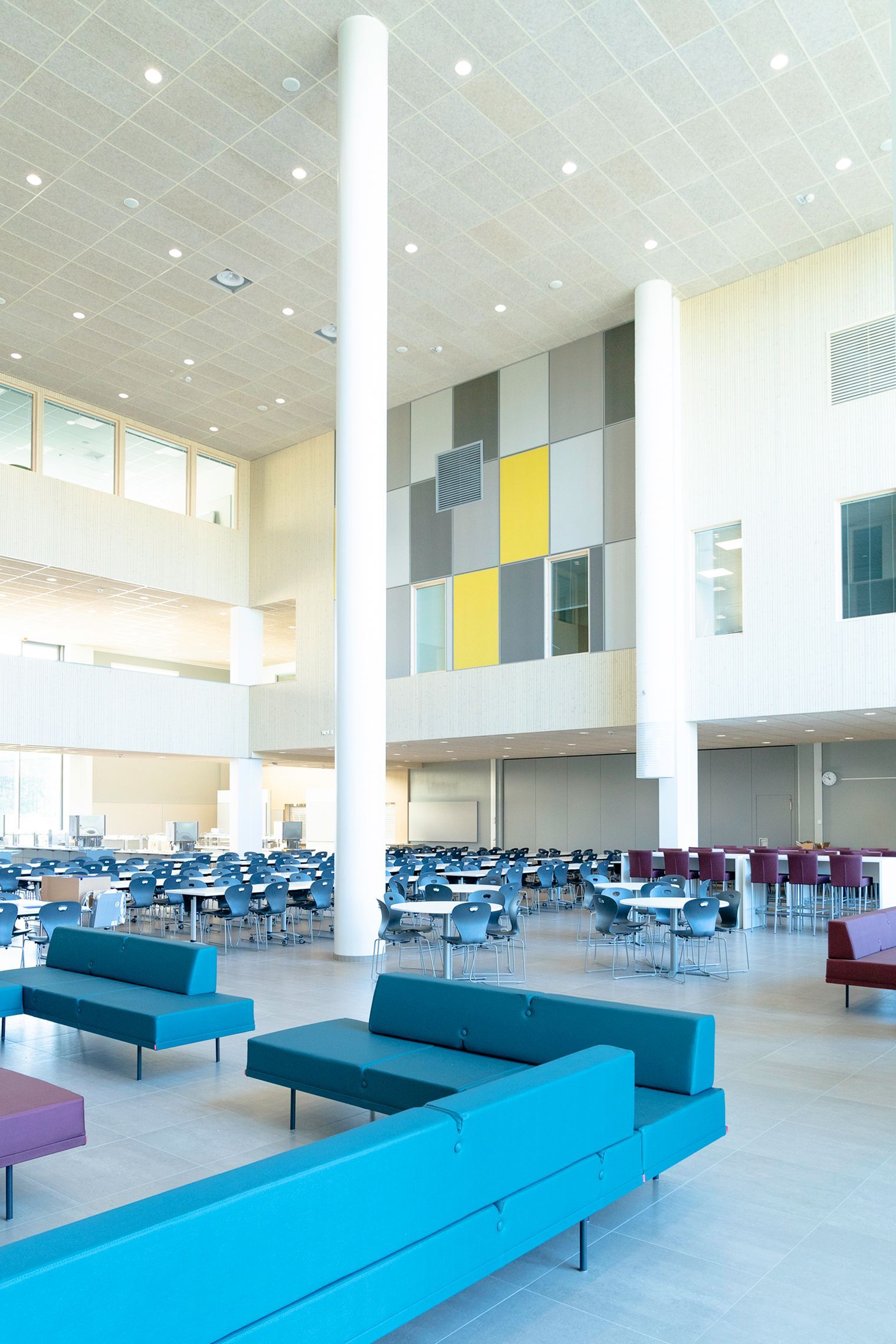 Romslig, moderne kantine. Blågrønne benker, bord med blå stoler, høy hvit søyle og fargerike veggpaneler.