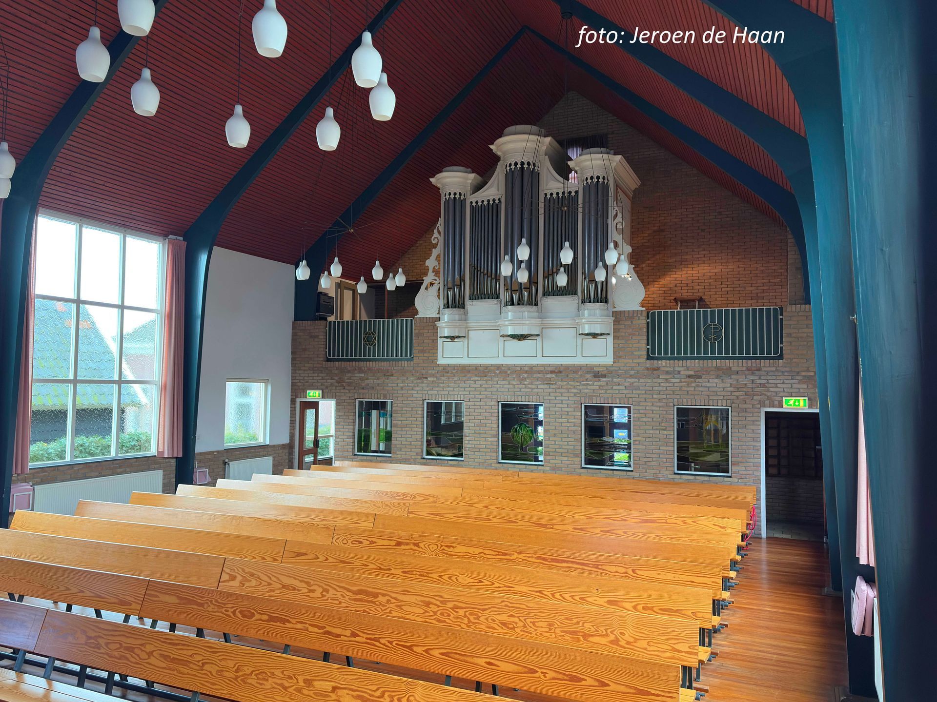 Interieur van een kerk met houten kerkbanken tegenover een orgel. Rood plafond, bakstenen muur en grote ramen.