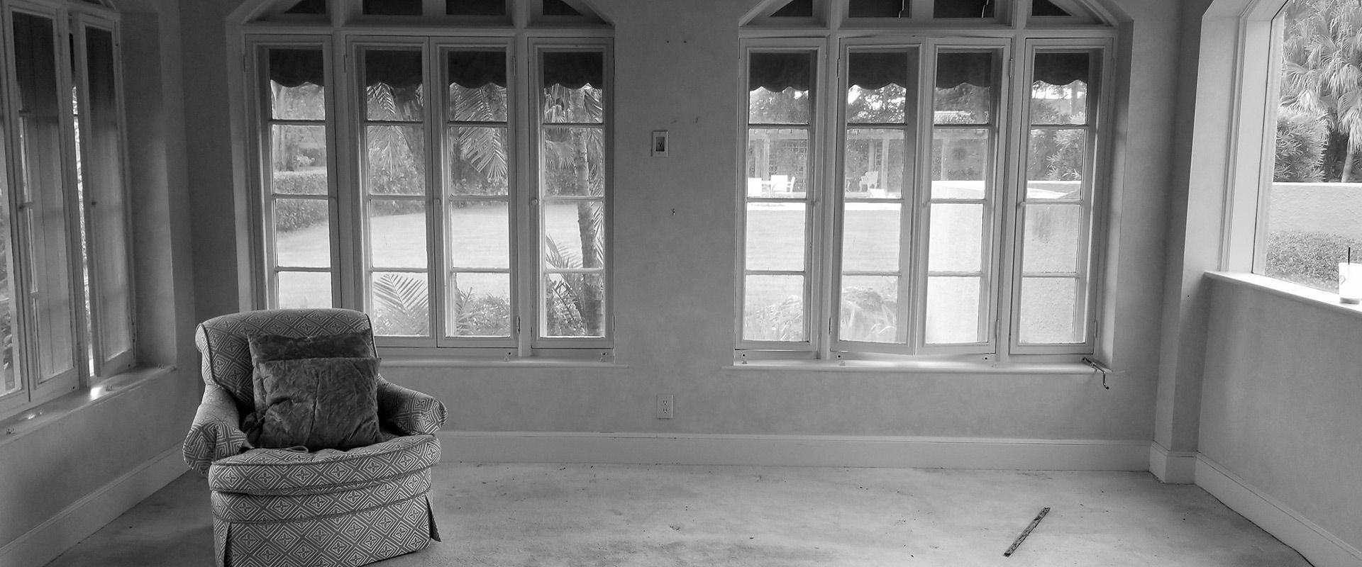 A black and white photo of an empty room with a chair and windows.