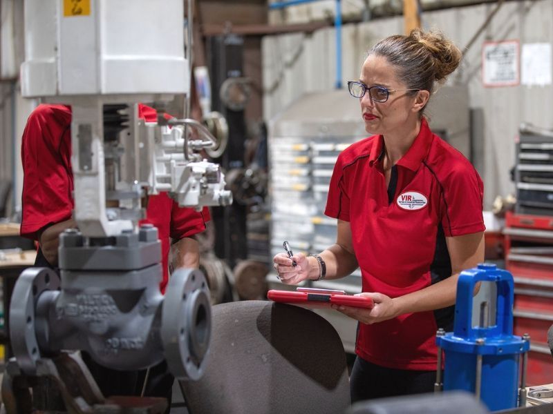 Worker in red shirt inspecting industrial piping with a clipboard in a factory