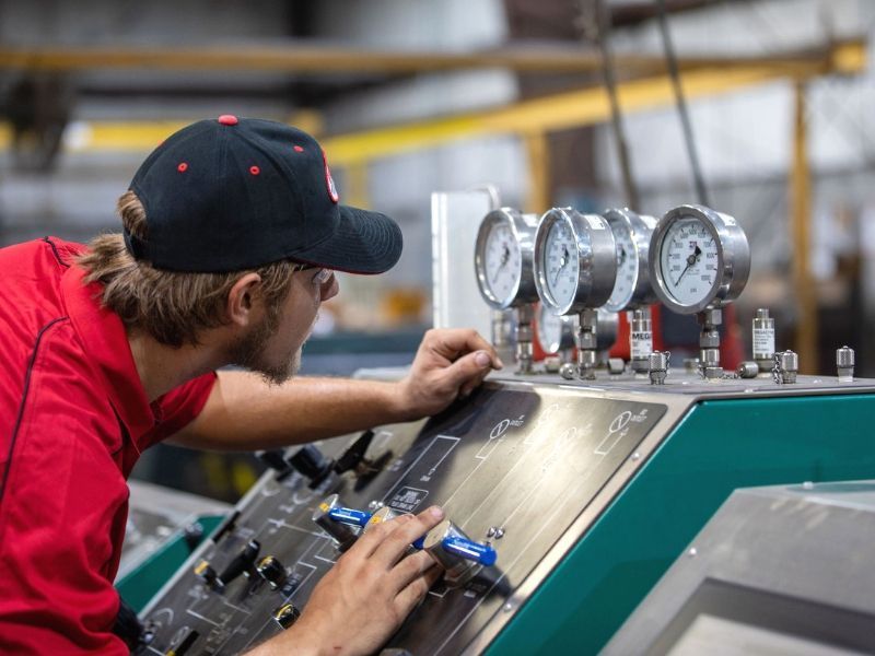 Technician in red shirt adjusts controls on industrial machinery with gauges in a workshop.
