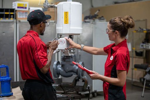 Two technicians in red uniforms inspect machinery in a workshop, one holding a tool and the other adjusting controls.