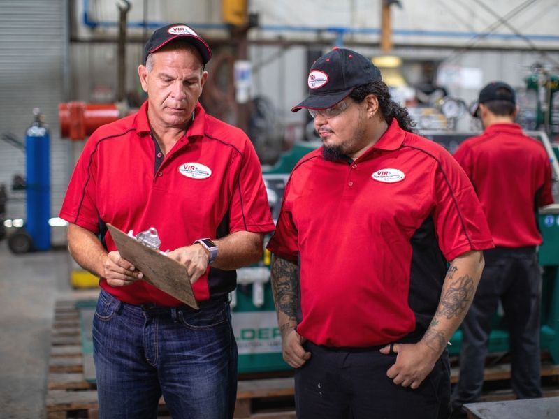 Two workers in red shirts review a clipboard in an industrial warehouse