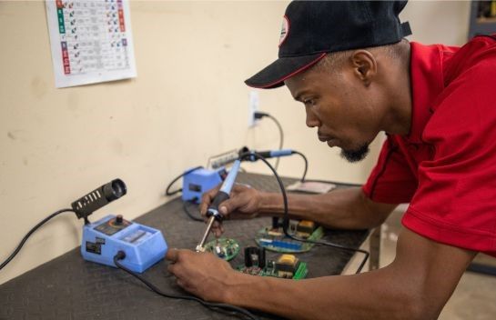 Technician in red shirt soldering a circuit board at a workbench with tools and electronics equipment