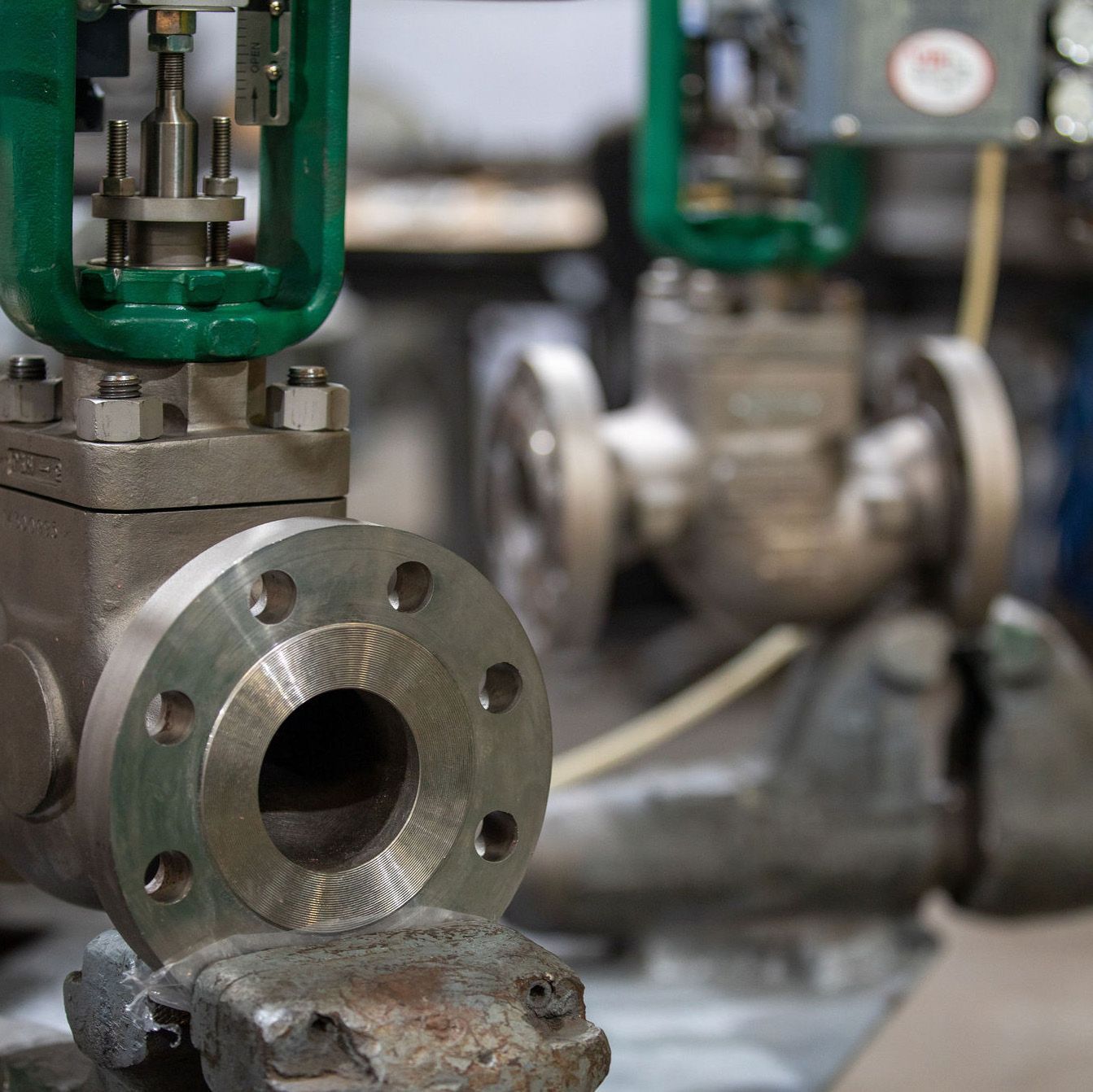 Industrial metal pipe flanges and valves in a factory setting, with a foreground pipe opening in focus