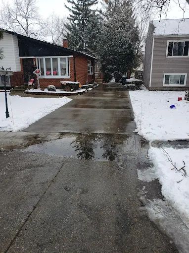 A snowy driveway with a puddle in the middle of it and a house in the background.