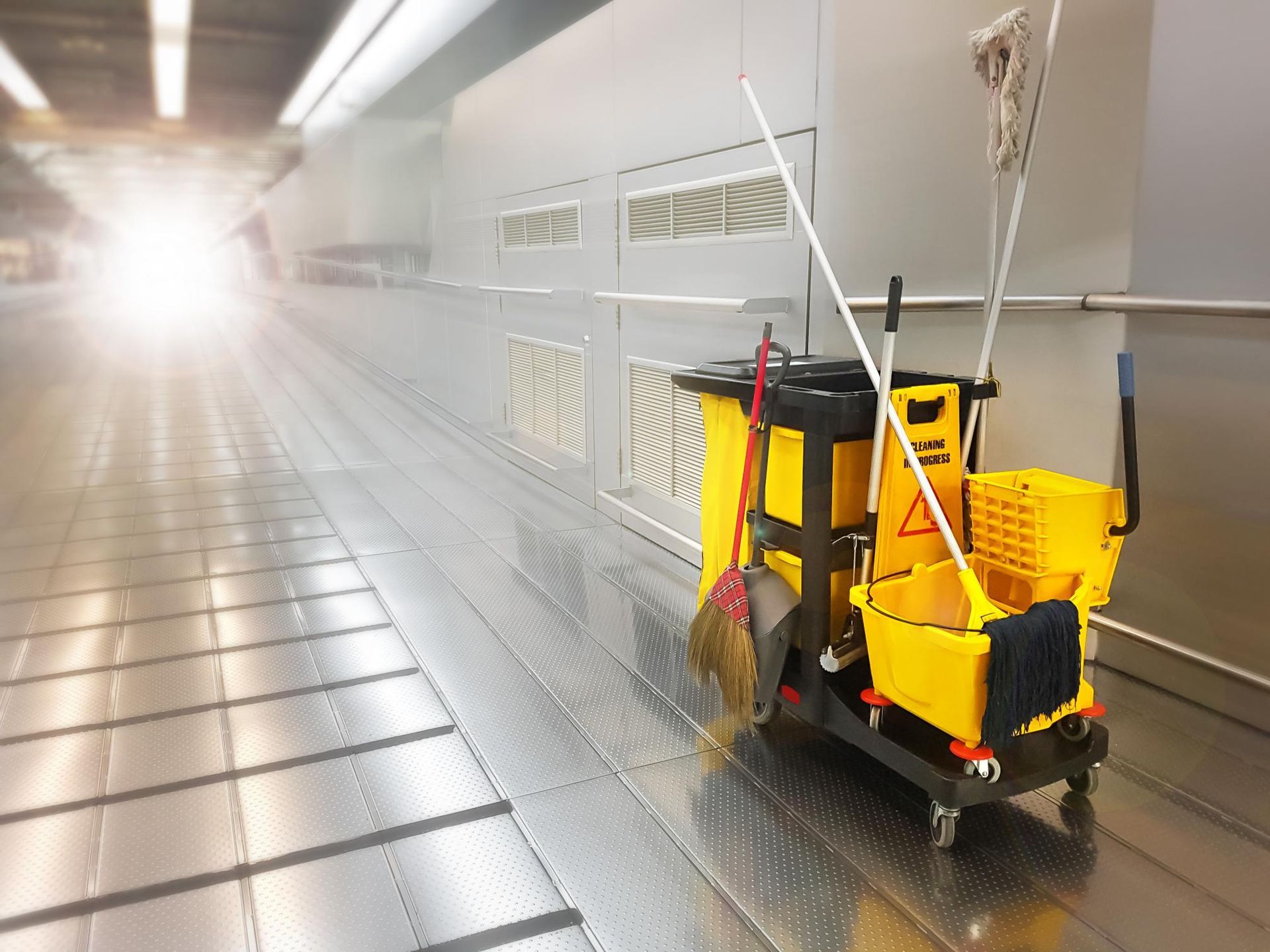 A yellow cleaning cart with a mop , broom and buckets in a hallway.