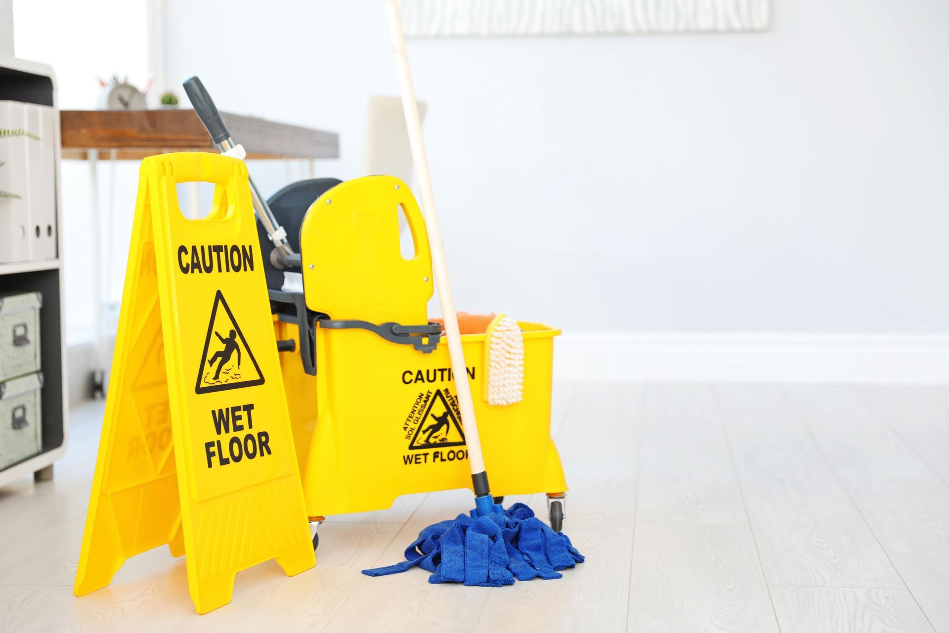 A wet floor sign , a mop , and a chair are on the floor in an office.