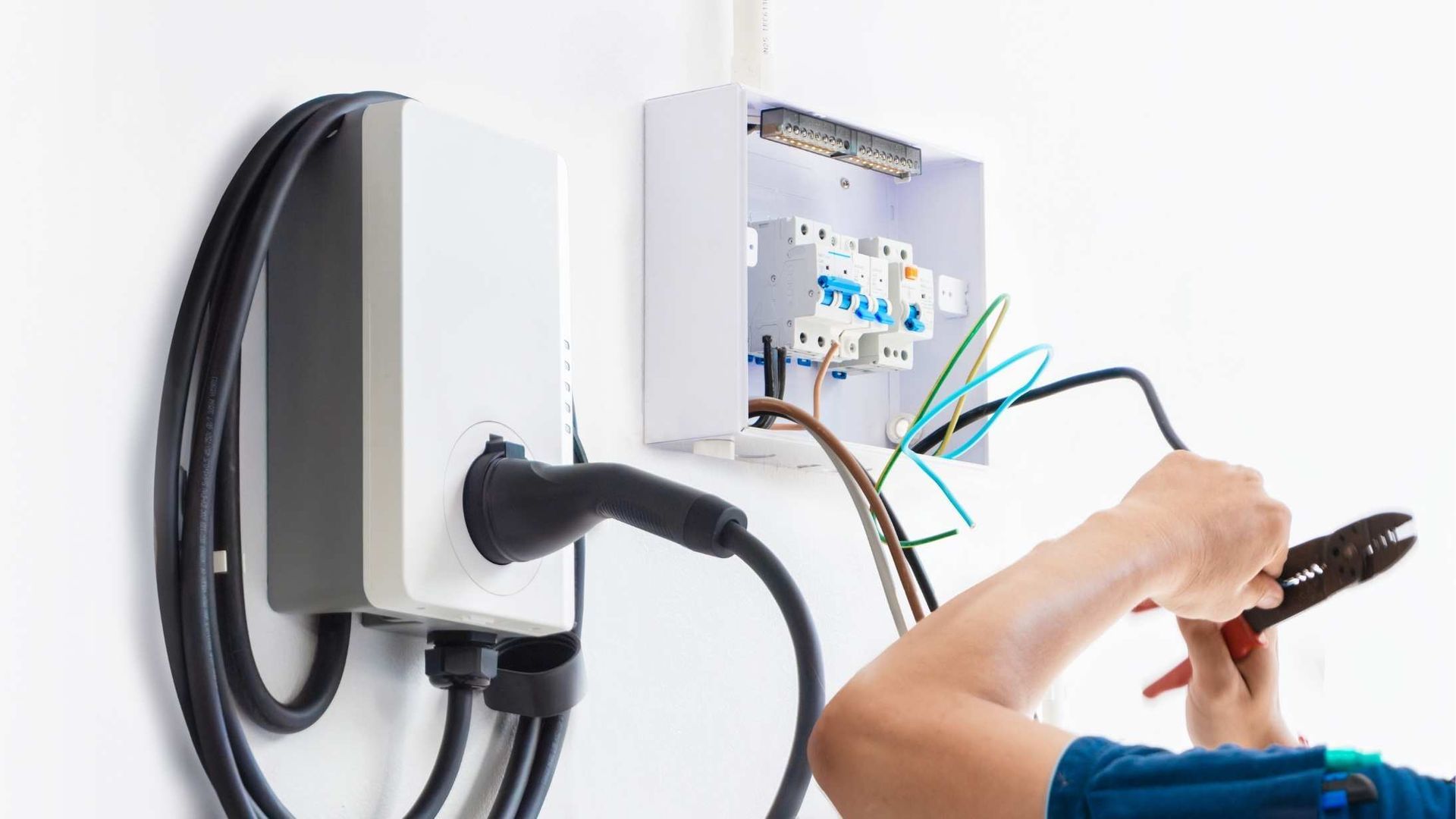 Electrician wiring an EV charger on a white wall, with the charger and electrical panel visible.