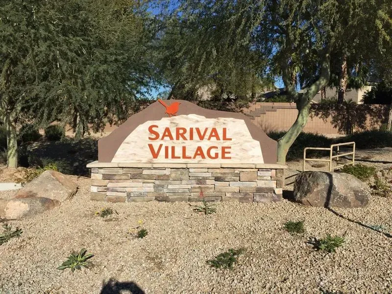 A sign for sarival village is surrounded by rocks and trees.