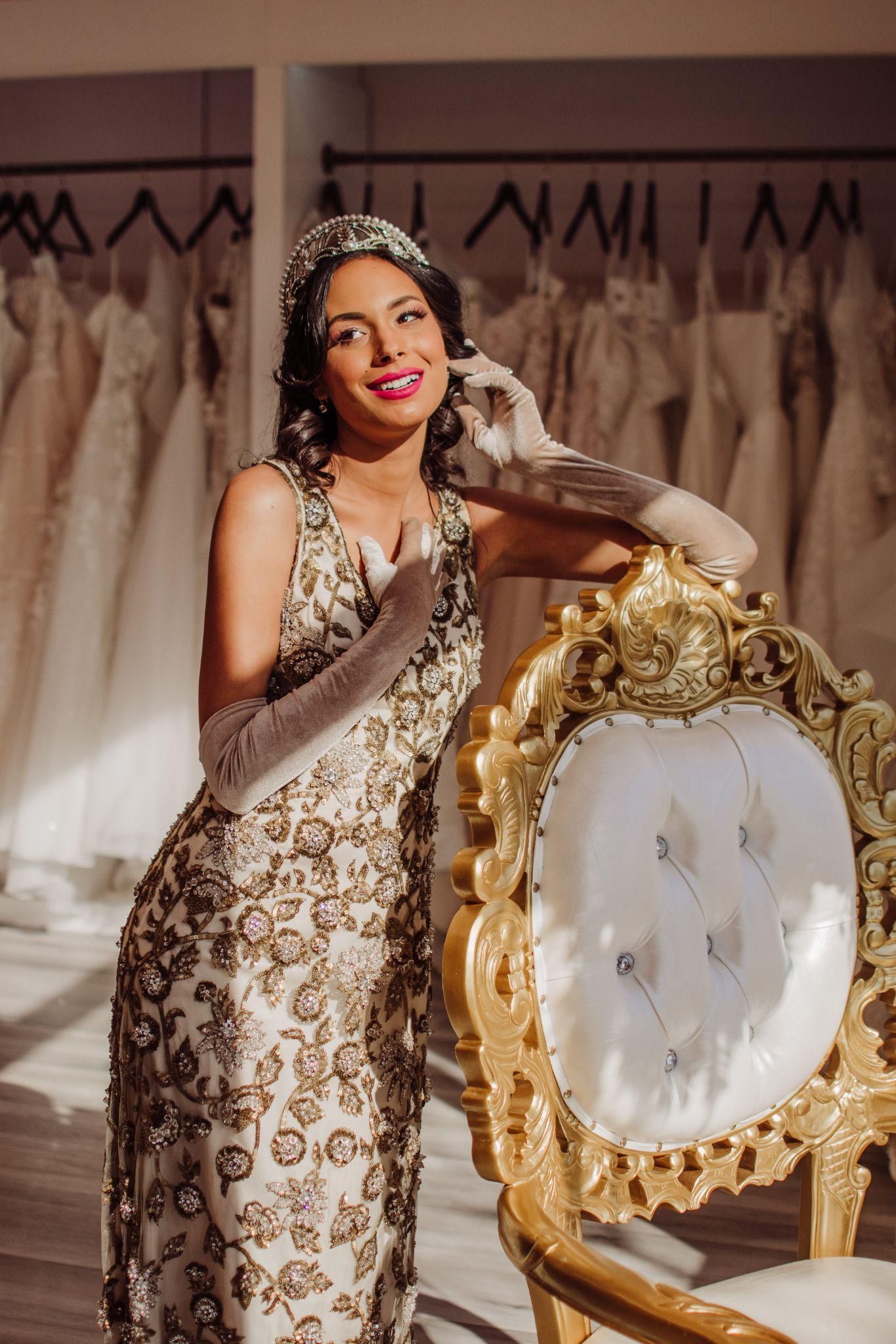 A woman in a wedding dress and gloves is standing next to a chair.