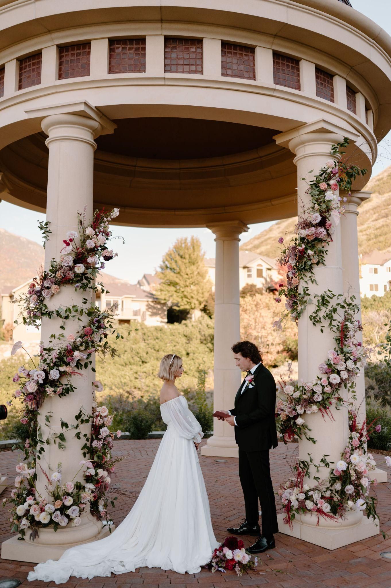 A bride and groom are standing under a gazebo decorated with flowers.