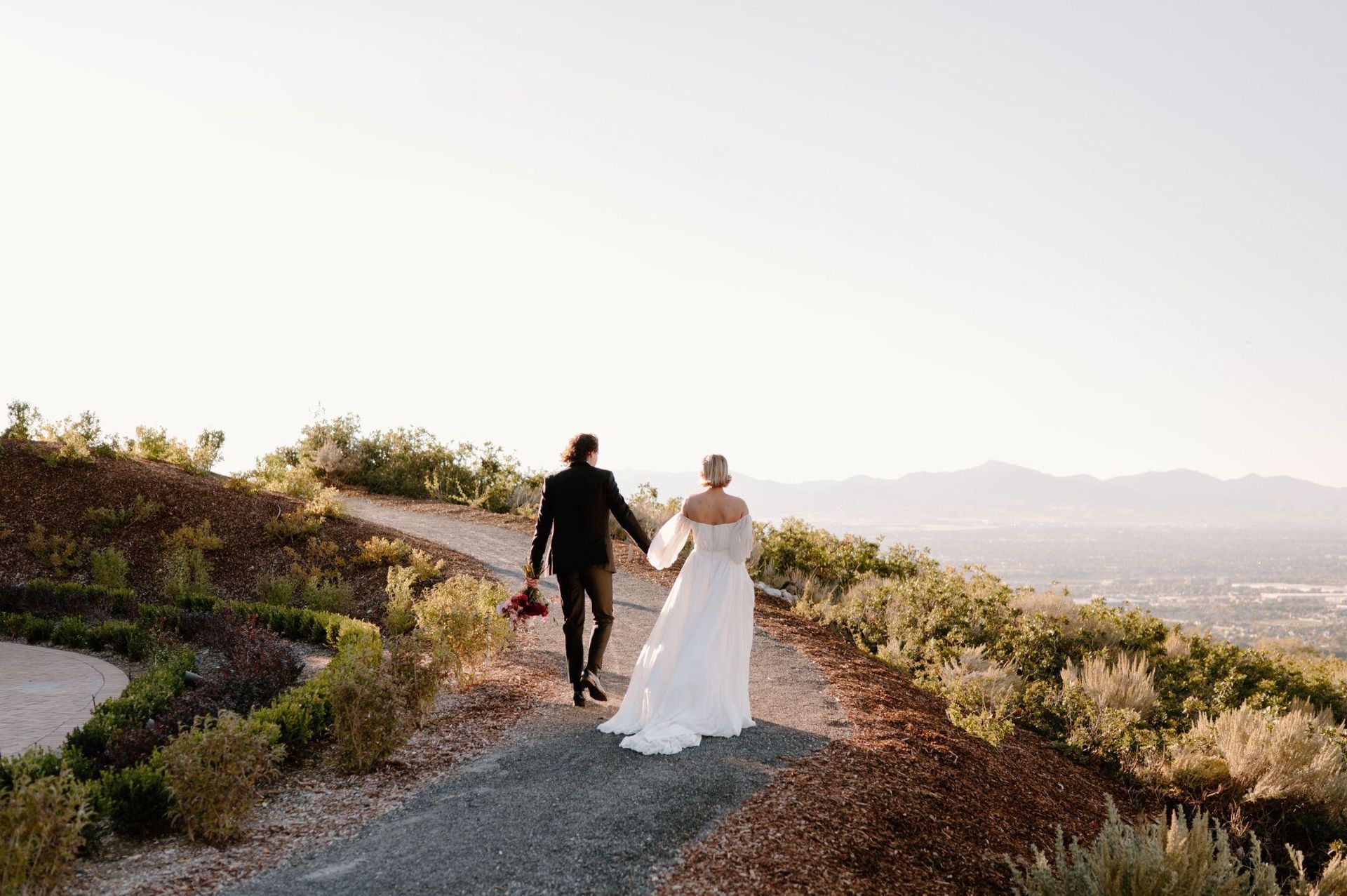 A bride and groom are walking down a dirt road holding hands.