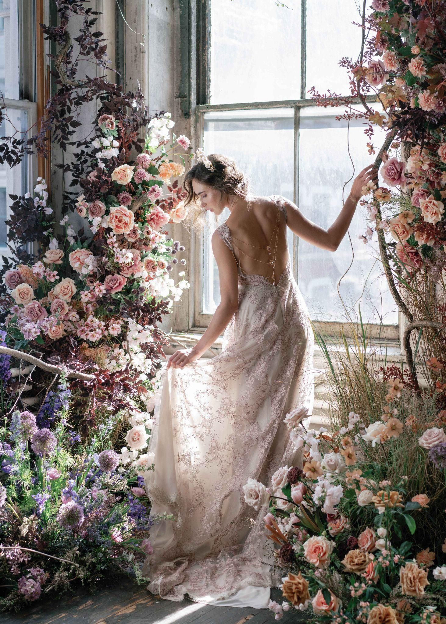 A woman in a wedding dress is standing in front of a window surrounded by flowers.