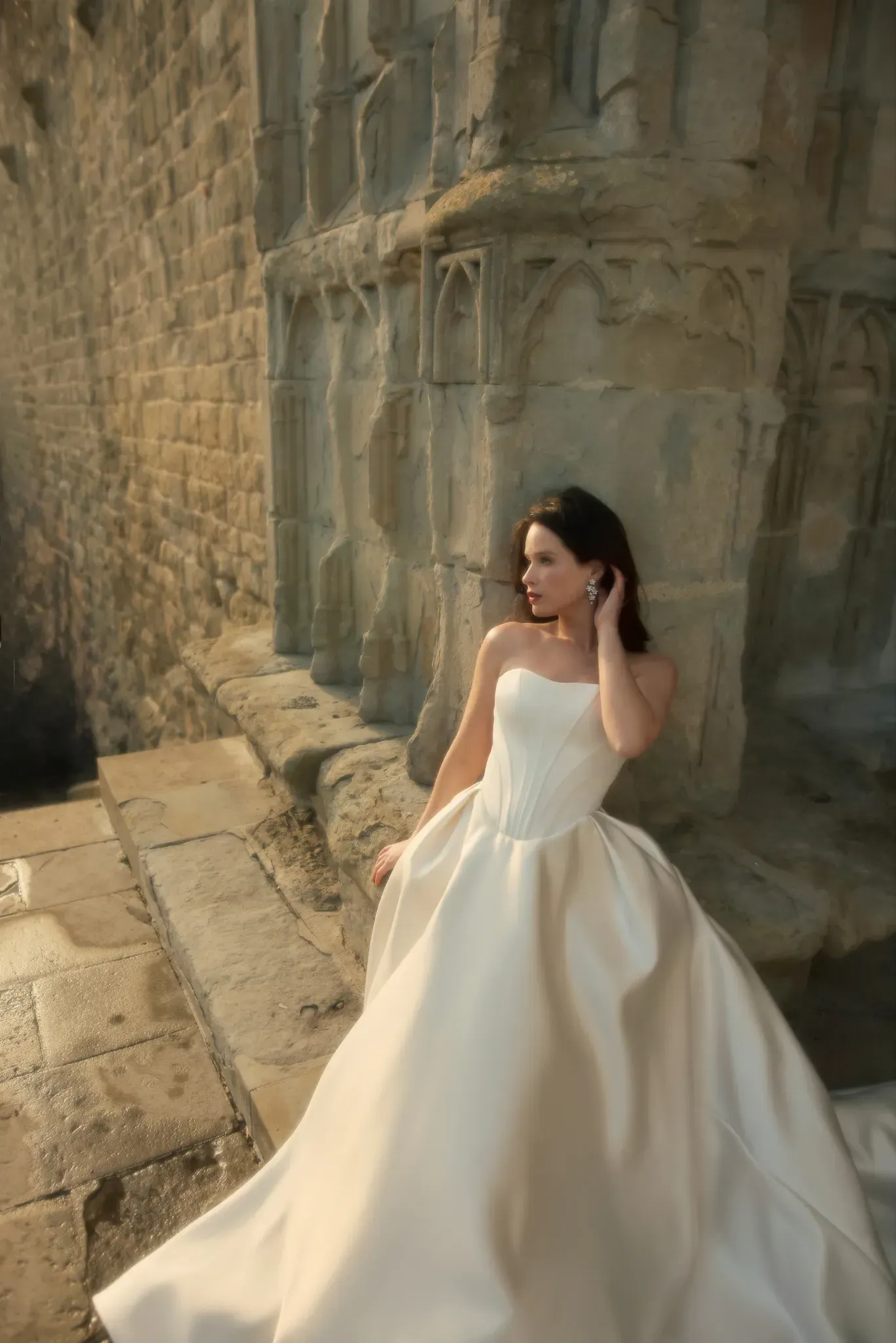 A woman in a white wedding dress is standing in front of a stone wall.