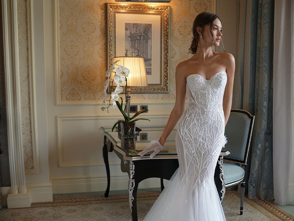 A woman in a lace wedding dress ang lace gloves is standing in a room.