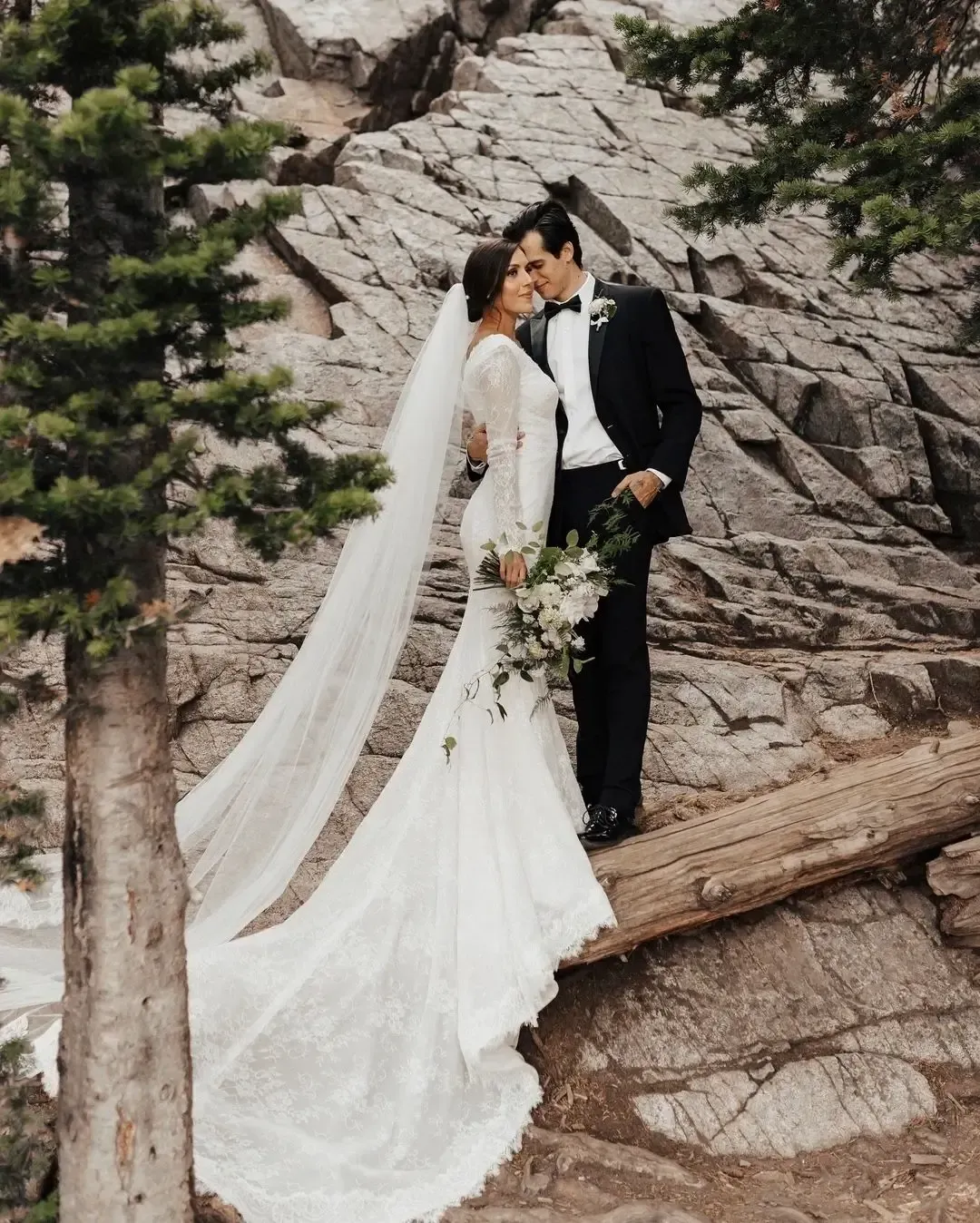 A bride and groom standing next to each other on a log
