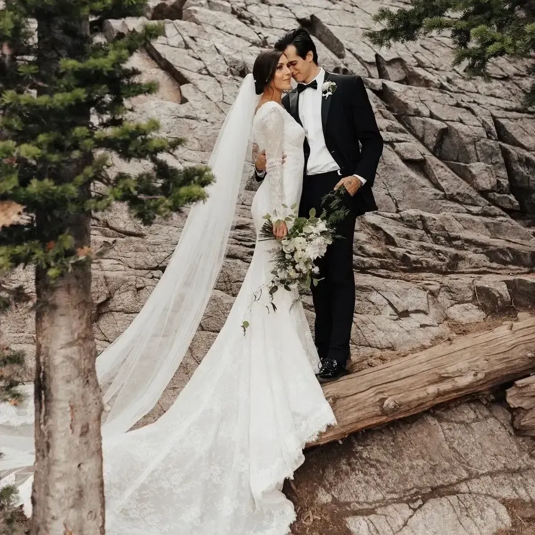 A modest bride wearing her custom wedding dress and her groom are posing for a picture on a log.