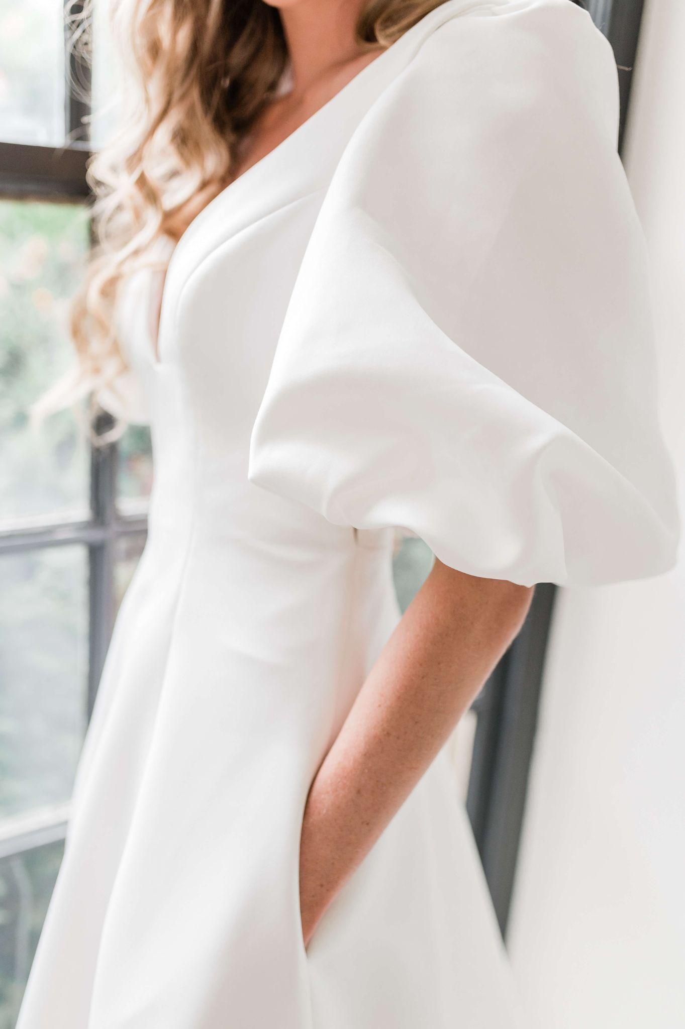 A woman in a white dress with puffed sleeves is standing in front of a window.