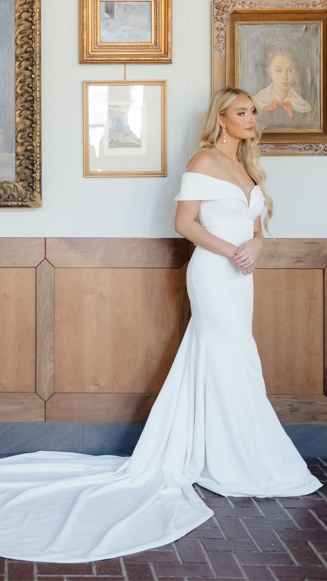 Woman in white off-the-shoulder wedding gown with long train, posed indoors near framed art.