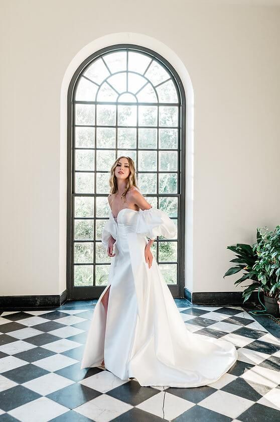 A woman in a wedding dress is standing in front of a window in a room with a checkered floor.