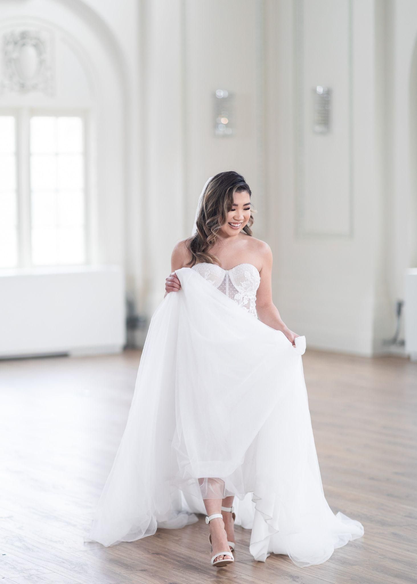 A bride in a white dress is standing in a room holding her dress.