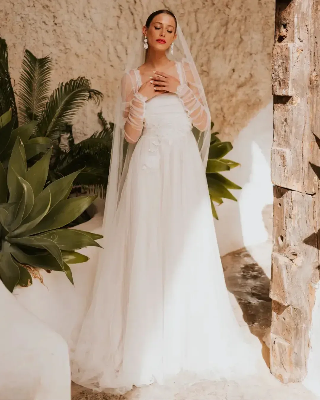 A woman in a white wedding dress and veil is standing in front of a stone wall.