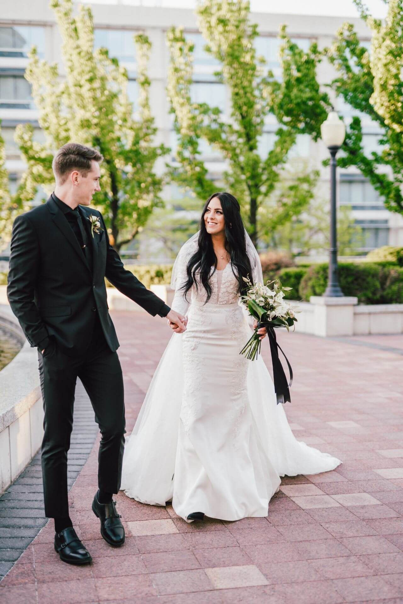 A bride and groom are walking down a sidewalk holding hands.