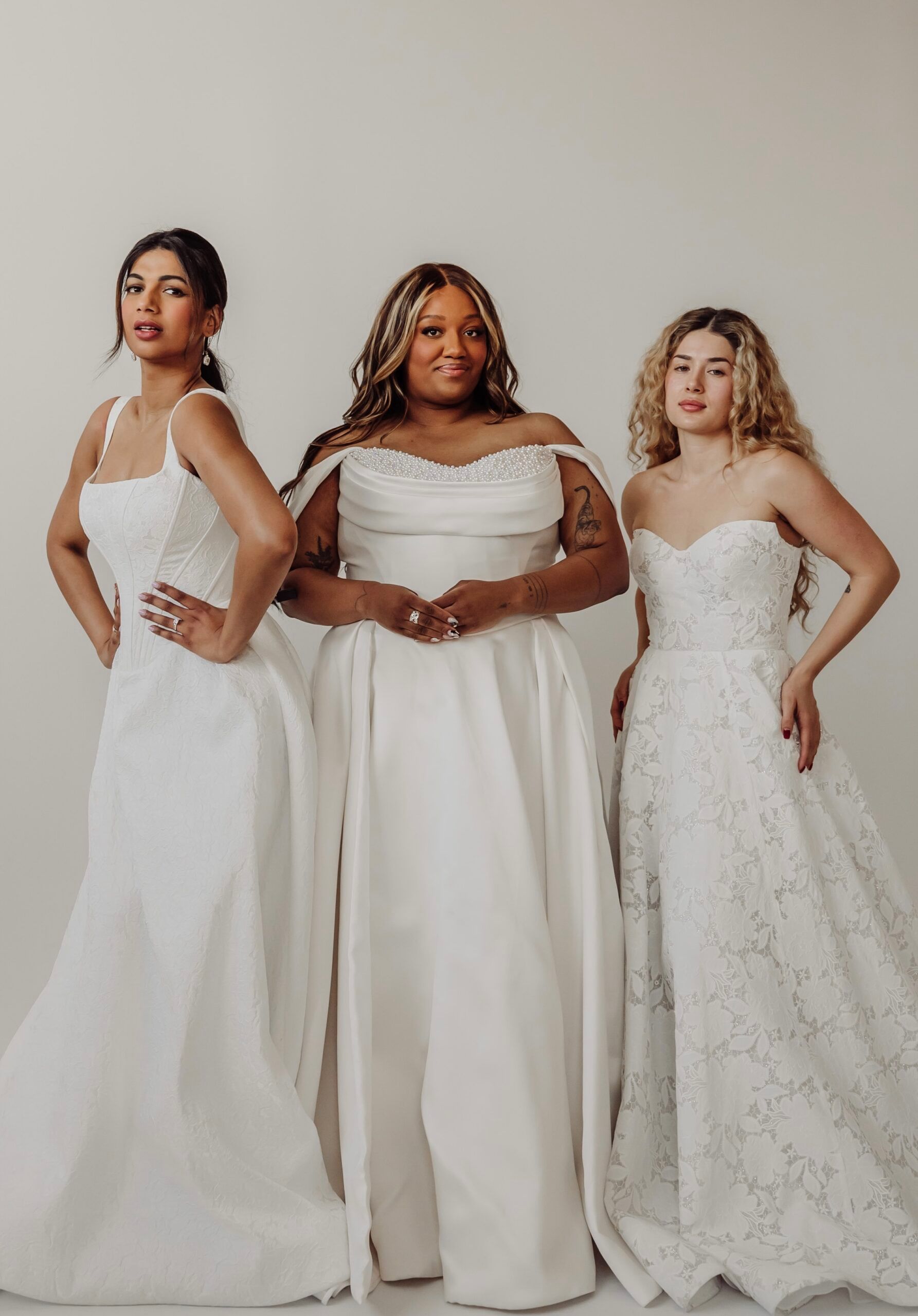 Three women of different sizes in white bridal gowns are standing next to each other.