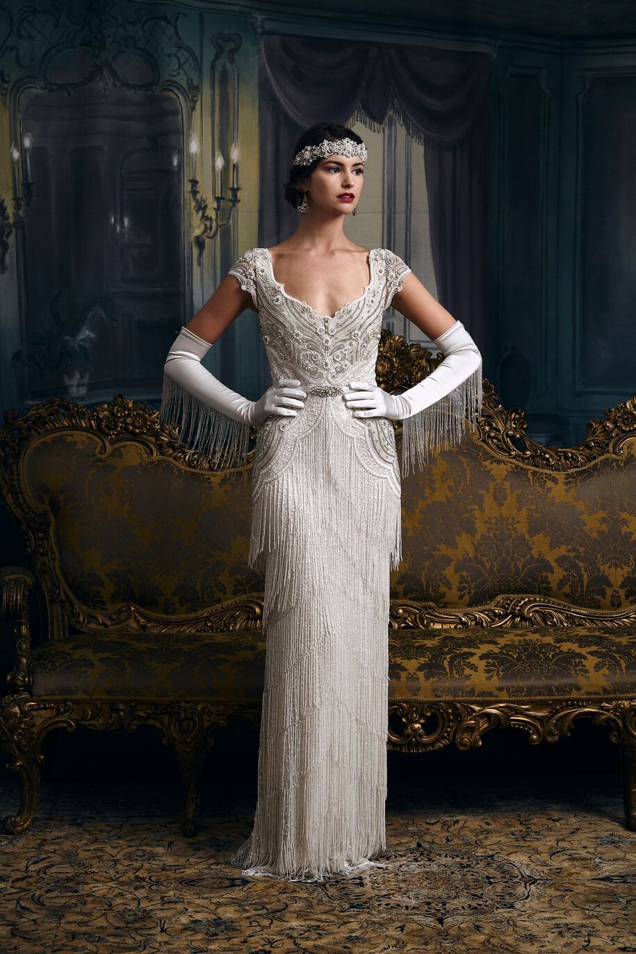 A woman in an Eliza Jane Howells' Carlotta wedding dress and gloves is standing in front of a couch.