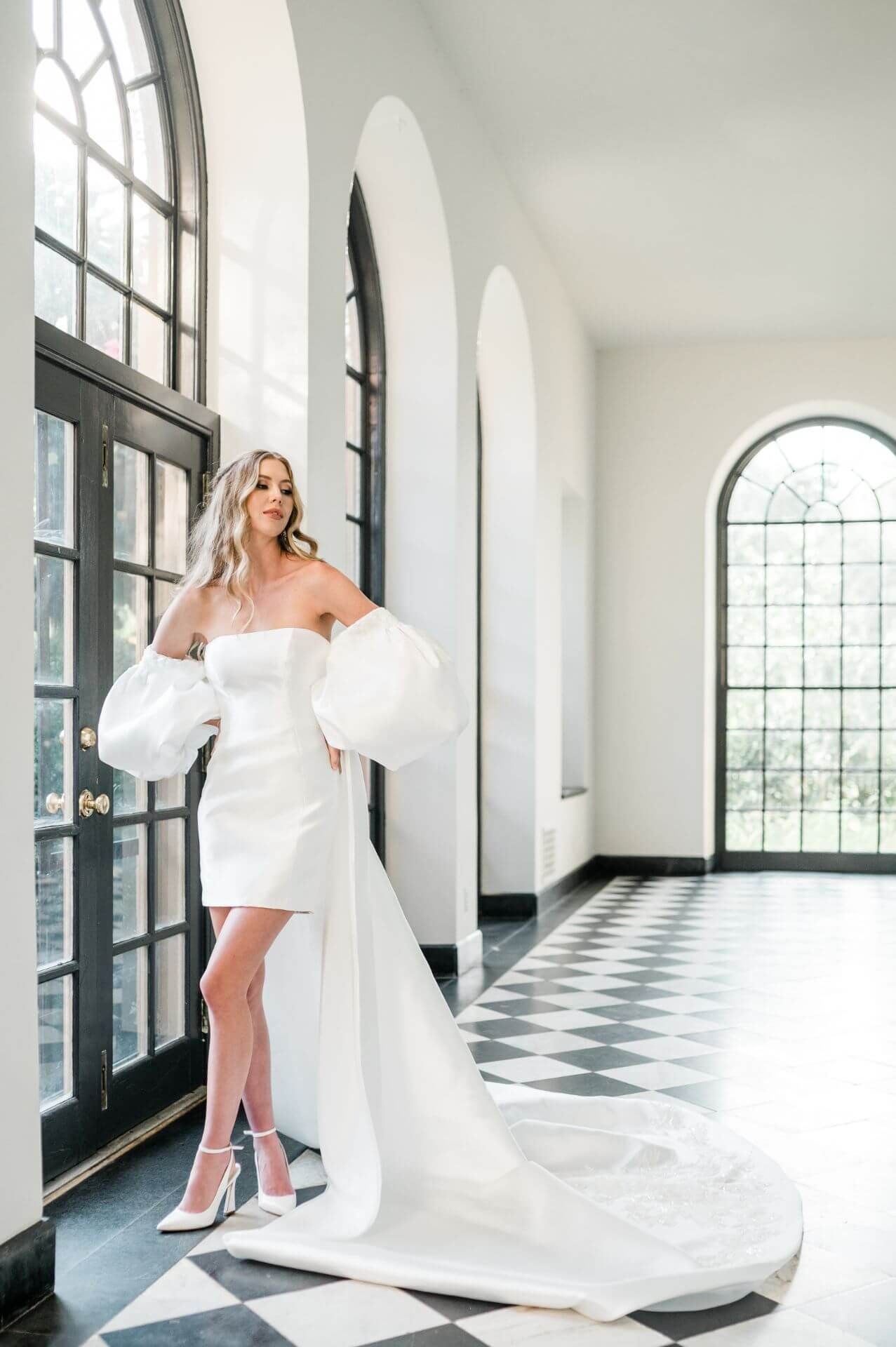 A woman in a white wedding mini-dress is standing in a hallway next to a window.