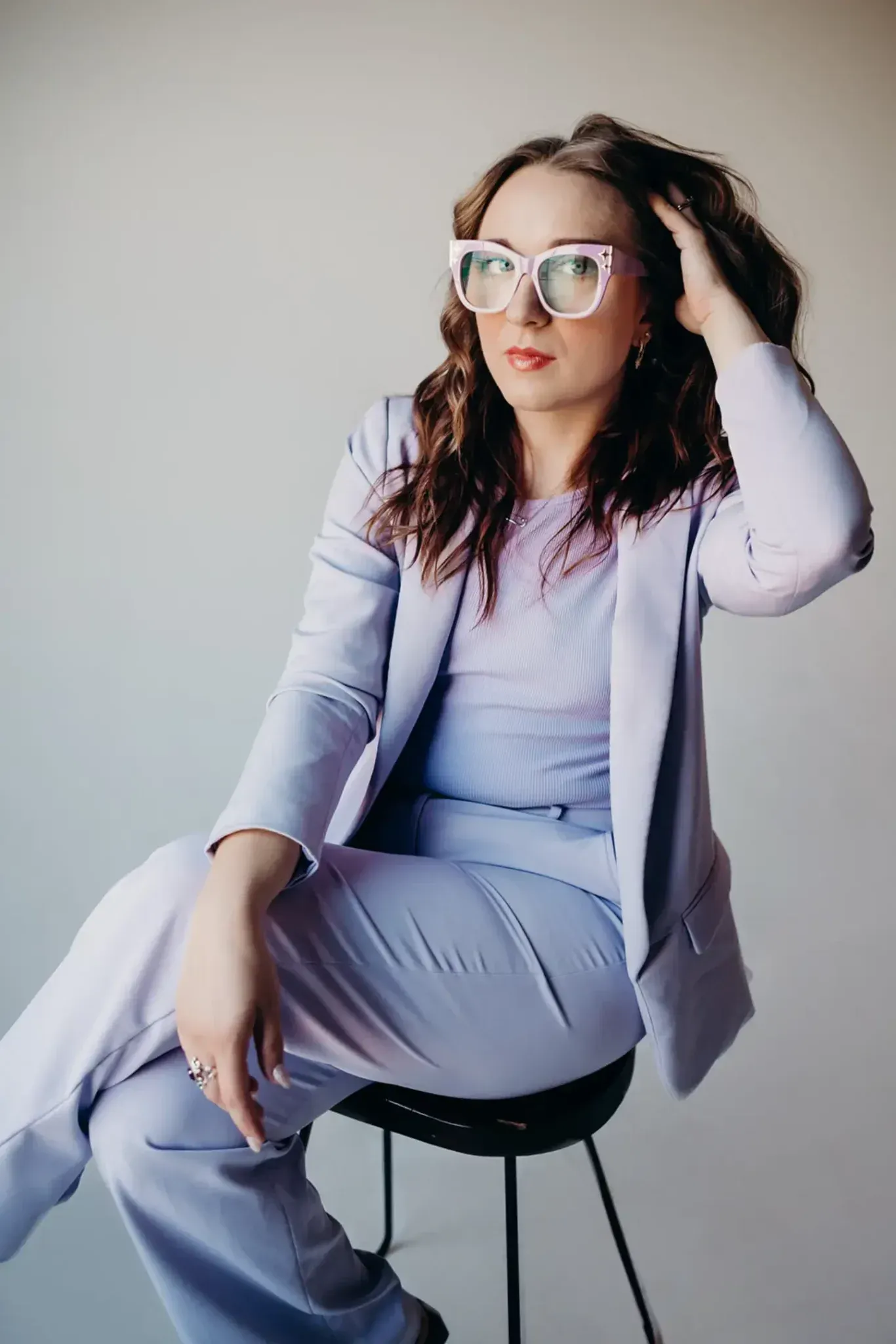A woman in a purple suit and glasses is sitting on a stool.
