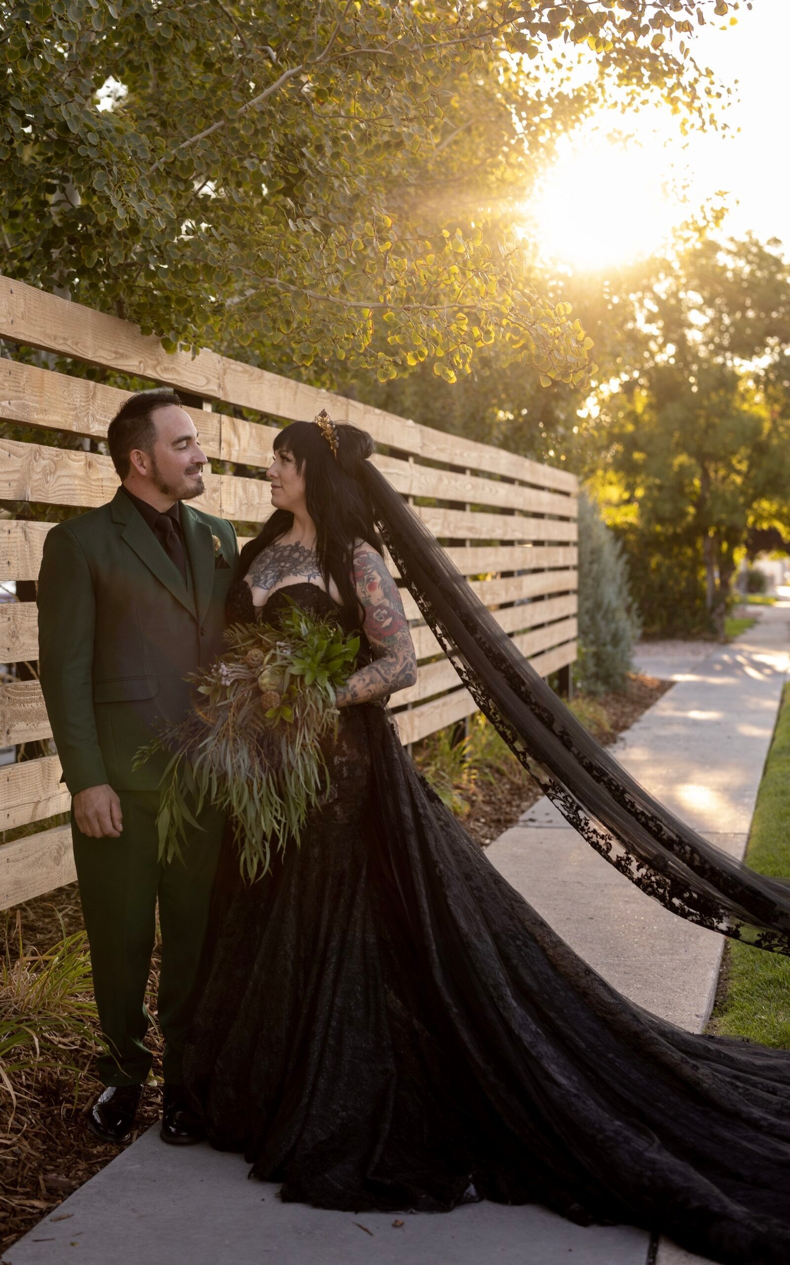 A bride in a black dress and a groom in a green suit are standing next to each other on a sidewalk.