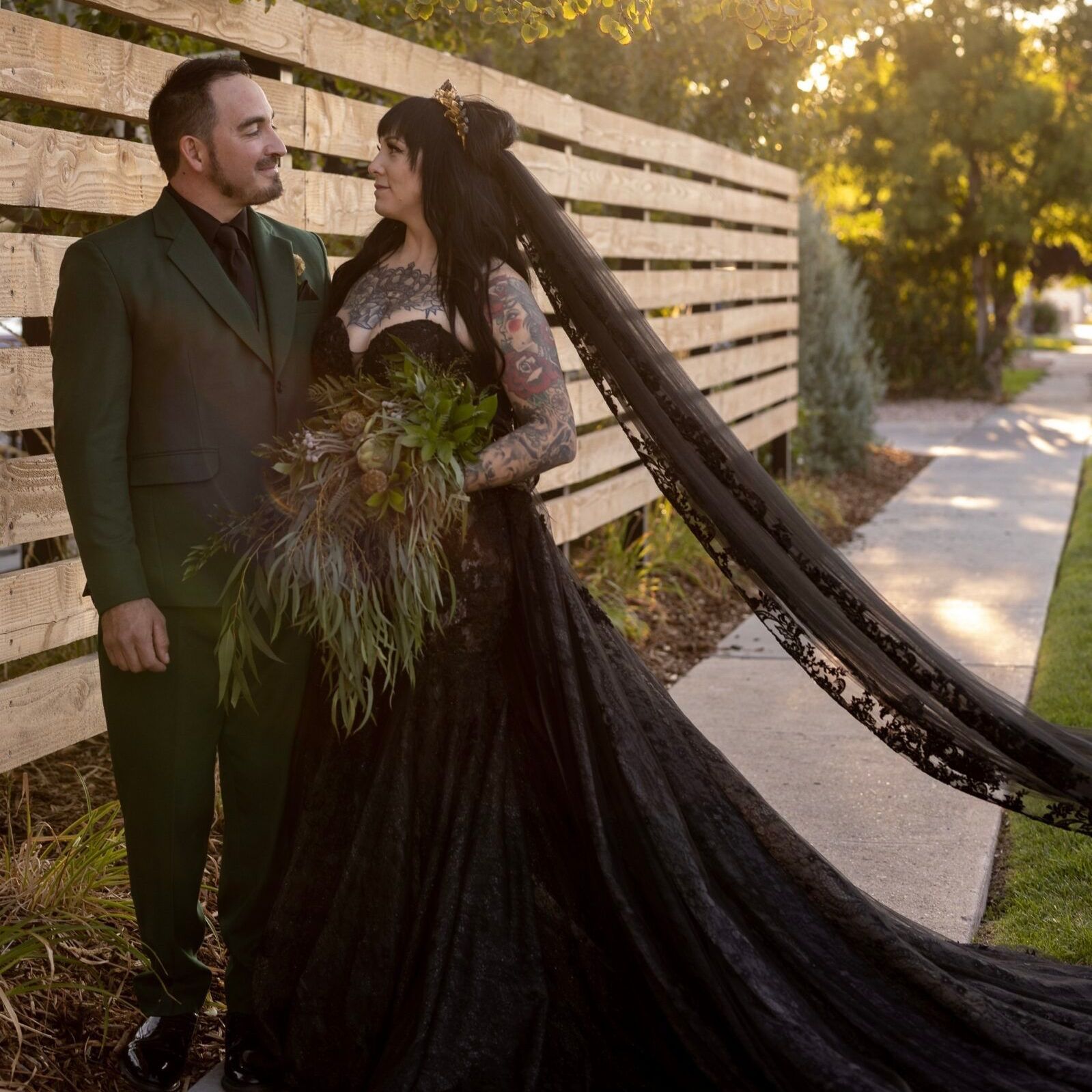 A man in a suit and a bride in a black wedding dress are standing next to each other on a sidewalk.