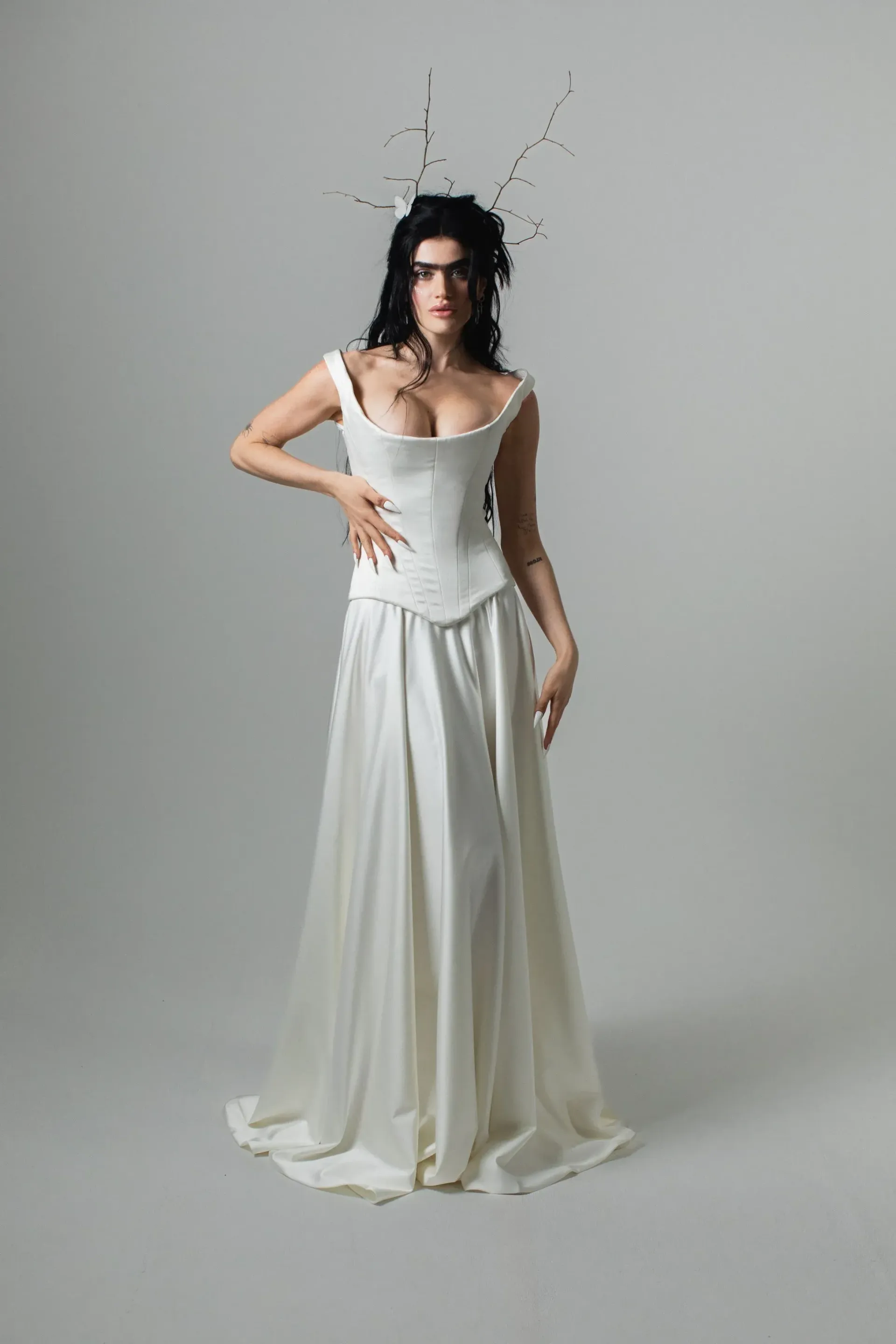 Hekate by Verander West Woman in white corset dress, dark hair, black headpiece, poses against light gray backdrop.