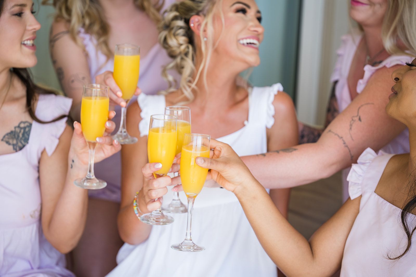 A Bride with her bridesmaids in lavender dresses, toasting with champagne cocktails, smiling, bright setting.