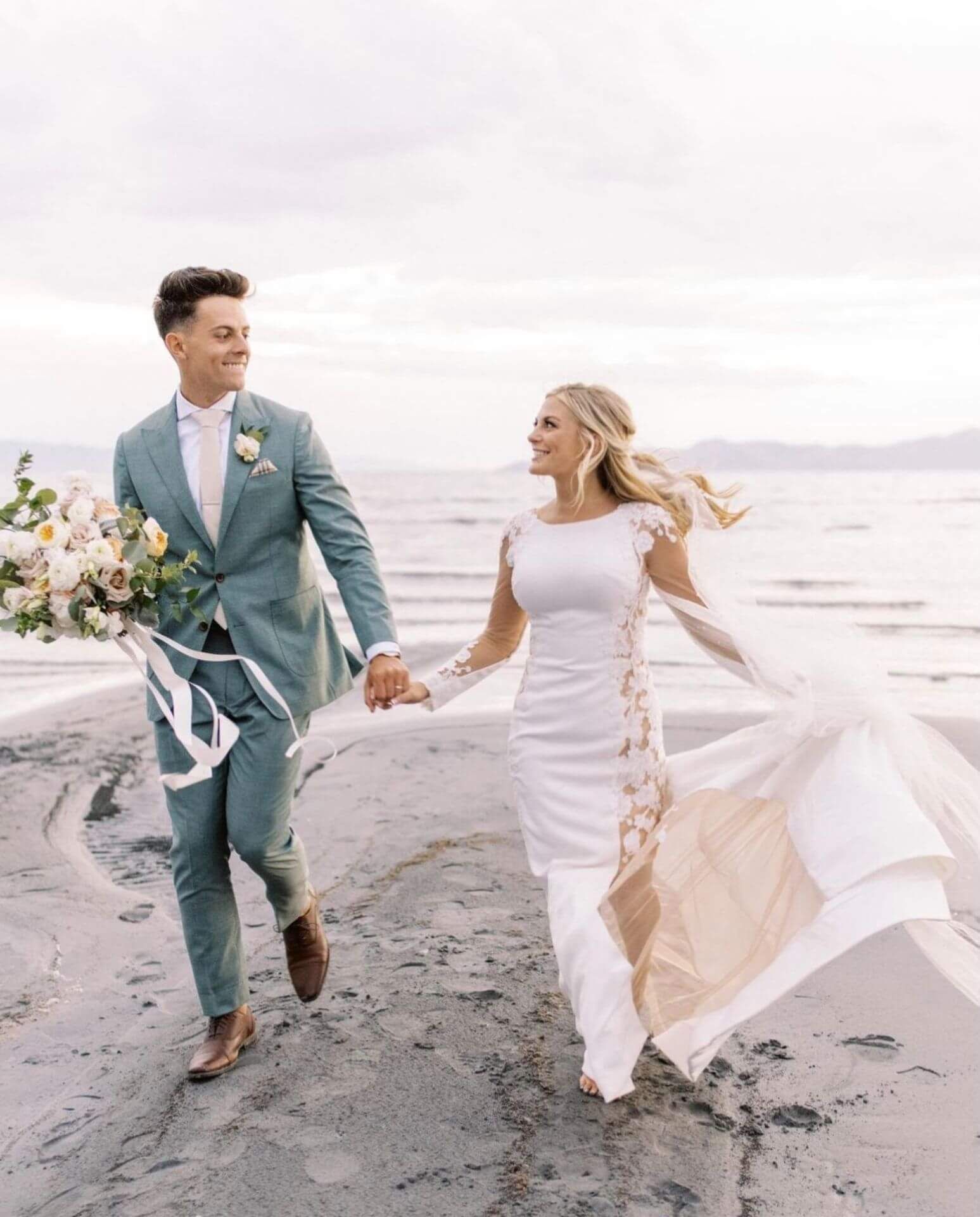 A bride and groom are walking on the beach holding hands.