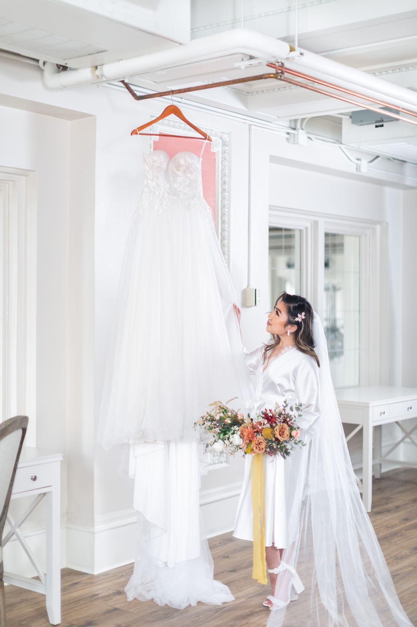 A bride in a white robe is holding a wedding dress hanging on a hanger.