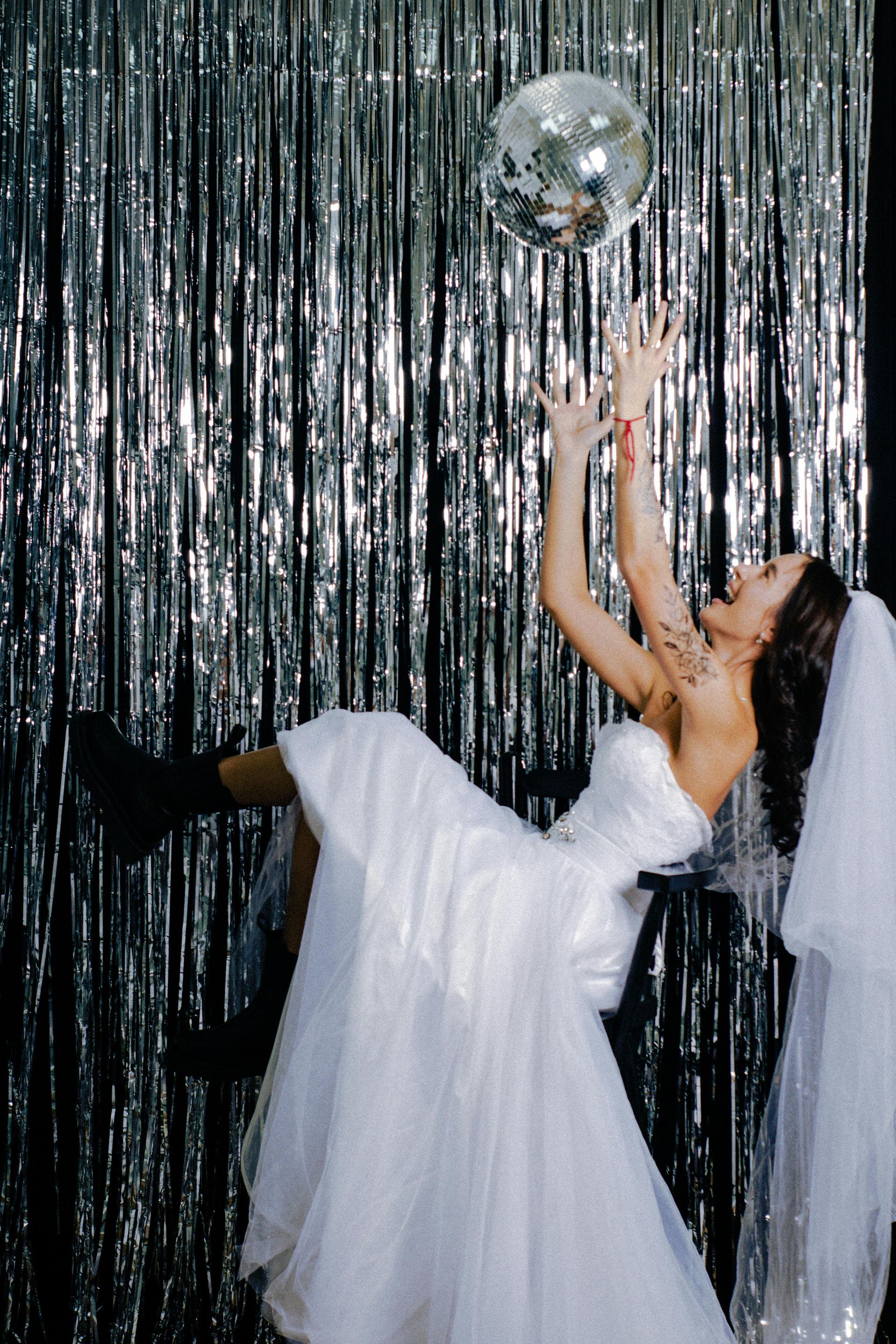 A woman in a wedding dress is reaching for a disco ball.