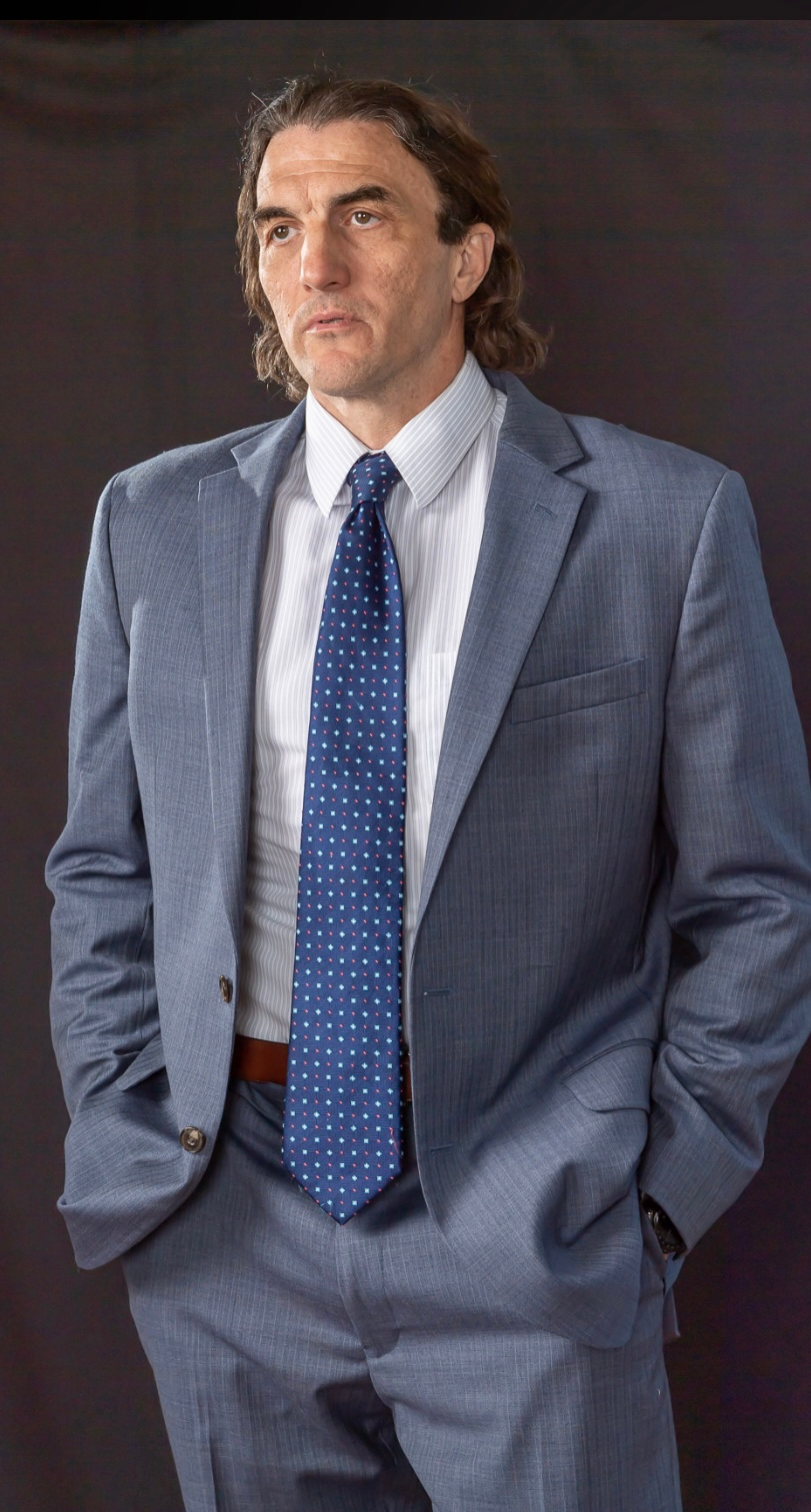 Man in a blue suit and tie, hands in pockets, looking off to the side.