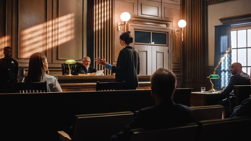 Woman speaking in a courtroom. Judge behind a desk. Sunlight streams through a window.