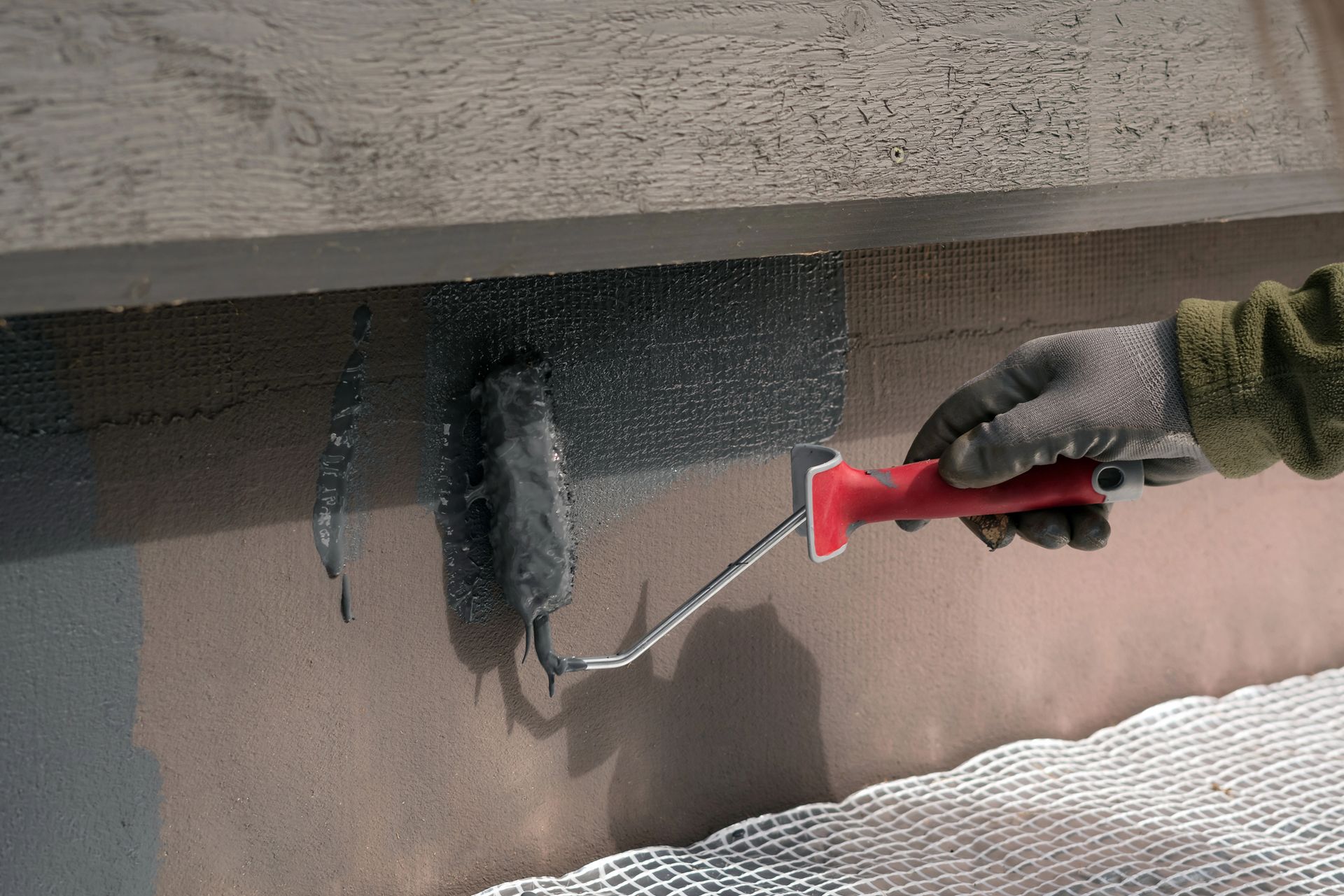 Persona che indossa guanti e applica una vernice scura sul muro con un rullo.